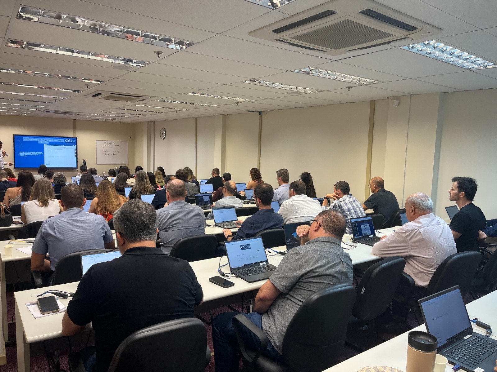 A foto mostra uma sala de treinamento cheia, com dezenas de pessoas sentadas em fileiras, todas utilizando notebooks. Na frente da sala, um instrutor apresenta o sistema Galileu em uma tela grande.