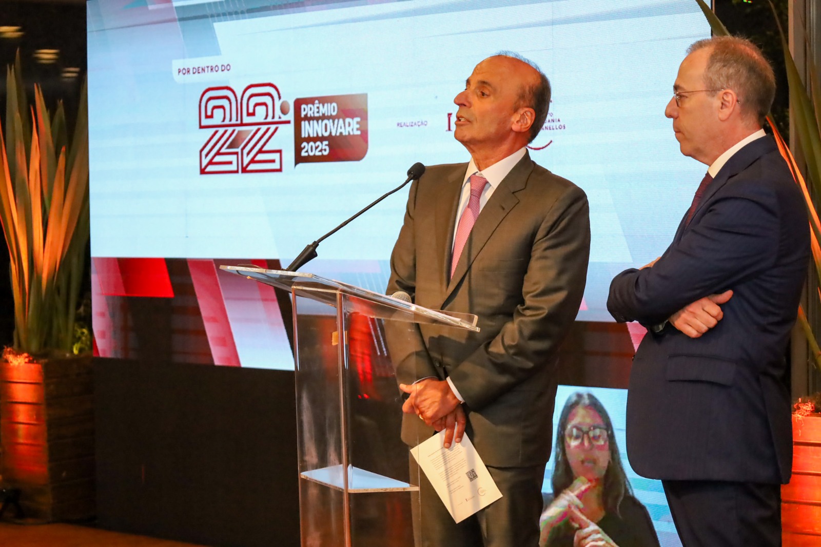A foto mostra os 2 representantes do Prêmio Innovare discursando num púlpito com um telão de fundo.