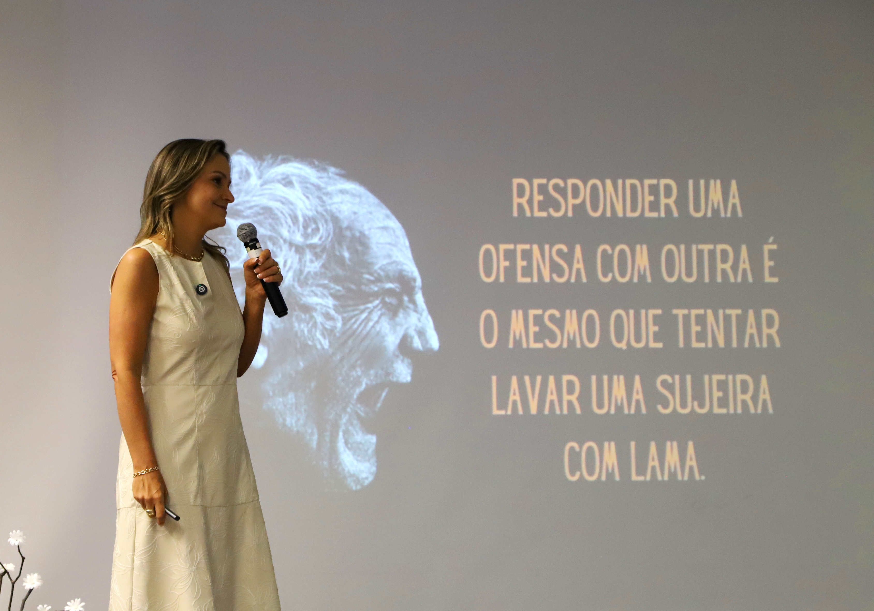 A fonoaudióloga e escritora Débora Brum fala ao microfone durante sua palestra no auditório Ruy Cirne Lima, com uma projeção ao fundo que mostra a frase: “Responder uma ofensa com outra é o mesmo que tentar lavar uma sujeira com lama.