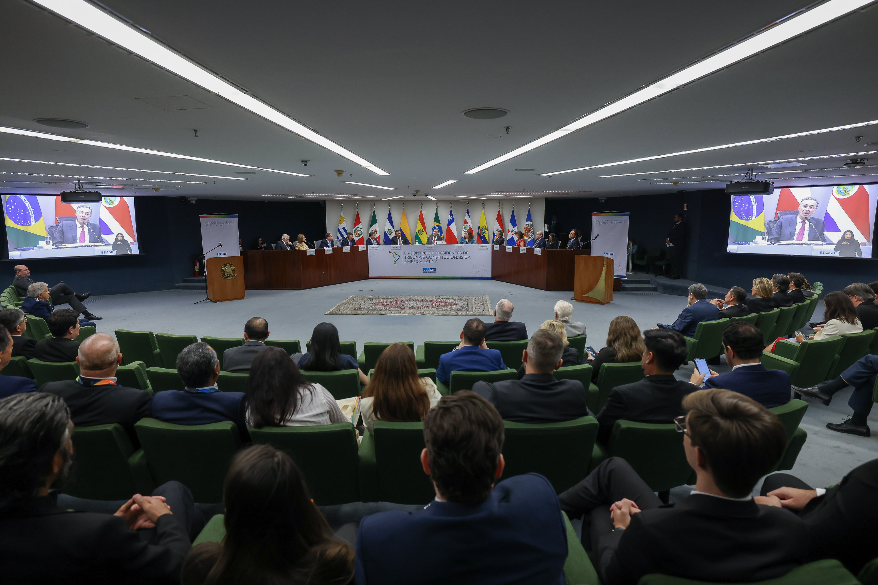 Auditório com diversas pessoas sentadas em poltronas verdes assistindo a uma conferência. Ao fundo, uma mesa composta por representantes de diferentes países, com bandeiras nacionais alinhadas atrás. Telões laterais mostram um orador discursando virtualmente, com as bandeiras do Brasil e da Costa Rica ao fundo. No centro, um painel indica que se trata do "Encontro dos Presidentes dos Tribunais Constitucionais da América Latina".