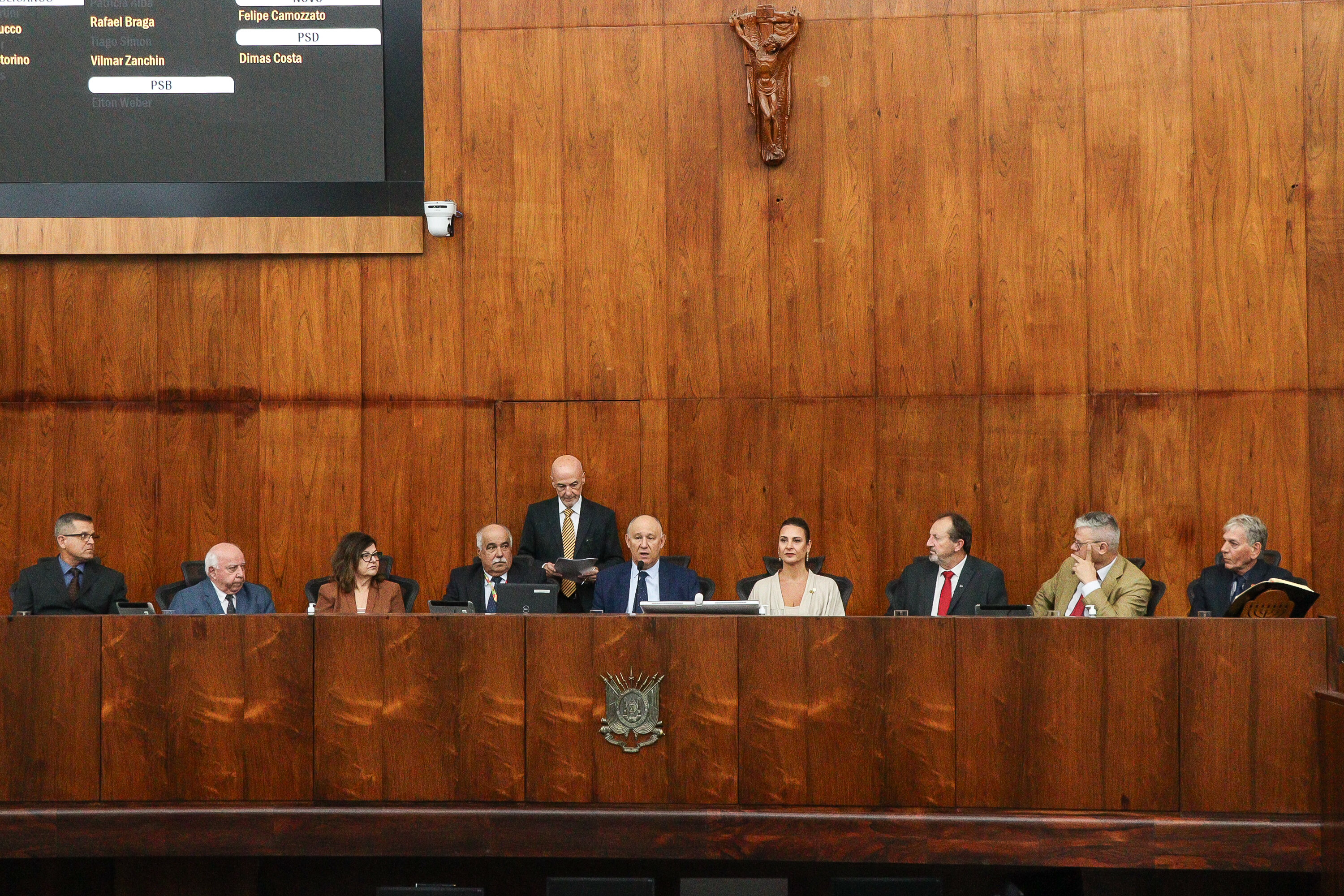 Sessão solene no Plenário 20 de Setembro da Assembleia Legislativa do Rio Grande do Sul. Nove autoridades estão sentadas na mesa diretora. A desembargadora de blazer marrom está entre os presentes, e um homem de pé lê um documento. O ambiente tem painel eletrônico de votação e parede revestida em madeira com crucifixo ao centro.