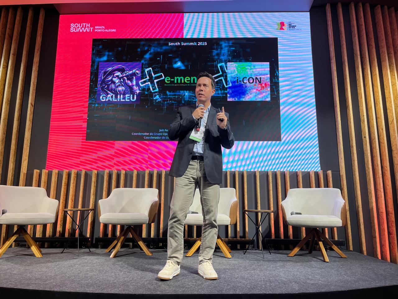 O juiz Rodrigo Trindade apresenta-se no centro do palco, diante de um telão vermelho e branco com o título "Avanços no Judiciário: Inteligência Artificial no TRT-RS", enquanto um fotógrafo registra o momento.