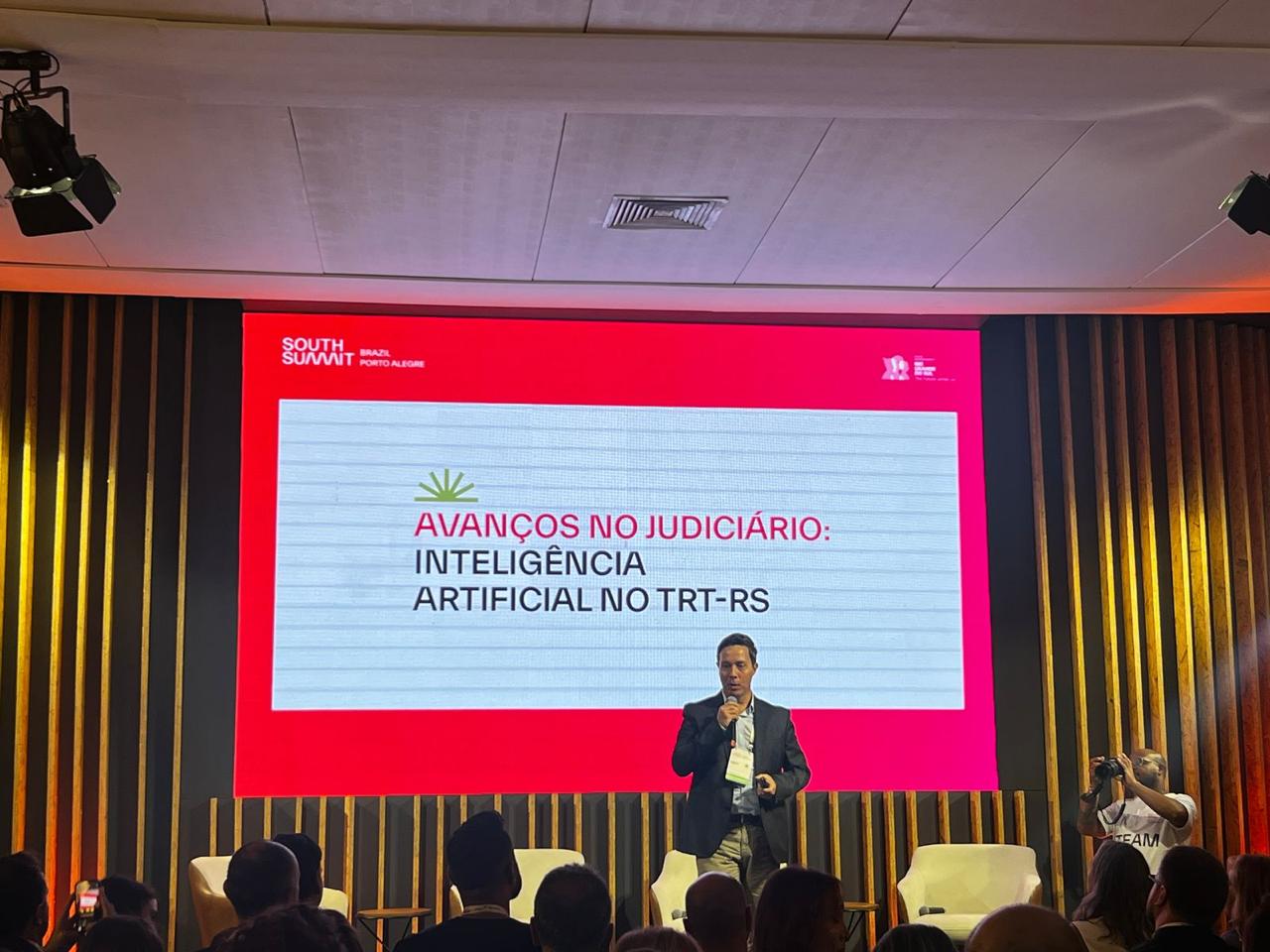 O juiz Rodrigo Trindade, de blazer escuro, camiseta branca e calça clara, discursa segurando um microfone em um palco com três poltronas claras e fundo de ripas de madeira; atrás dele, um telão exibe artes digitais dos projetos Galileu, e-mentores e i-CON.