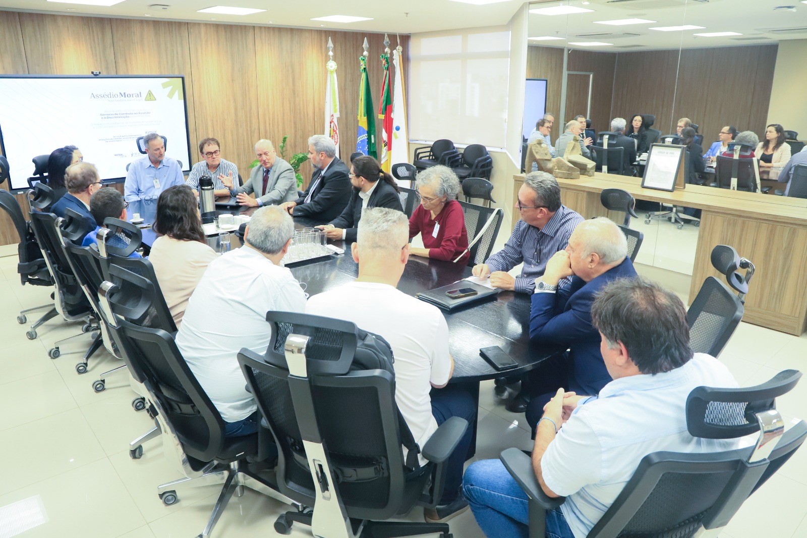 Participantes na mesa de reunião. Martins Costa aparece em destaque falando e gesticulando. Ao fundo aparecem bandeiras e um telão ligado.