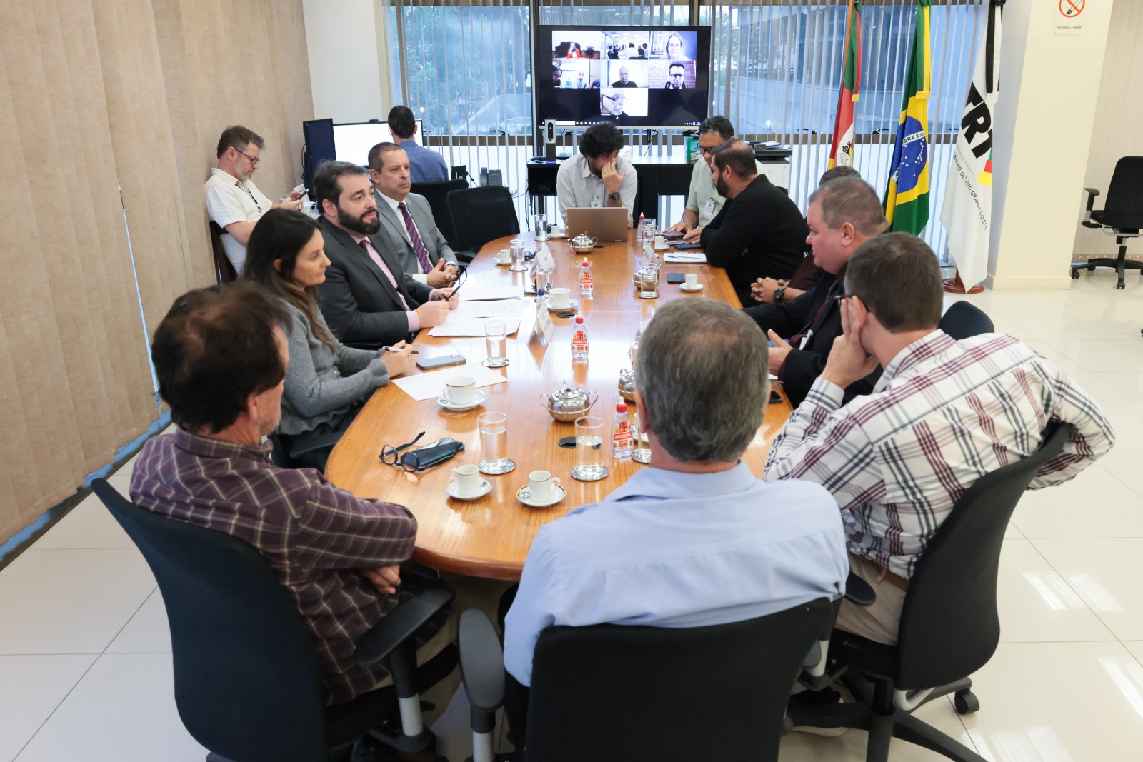 Pessoas na mesa de negociação. Aparecem 13 pessoas. Ao fundo, um telão ligado e as bandeiras do RS, do Brasil e do TRT-RS.