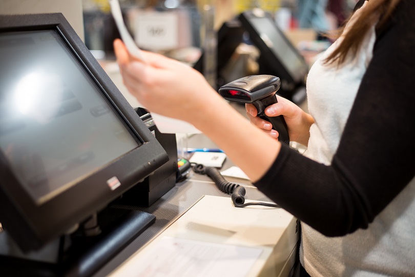 Foto ilustrativa mostra trabalhadora, caixa de supermercado, segurando equipamento leitor de código de barras. Ela está de costas, tem o cabelo preto e comprido. Veste blusa preta e colete branco.