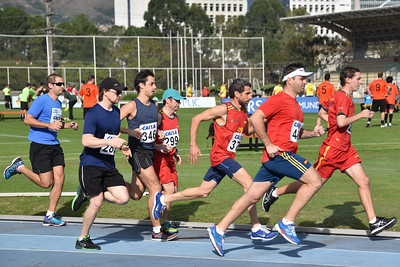 Sete pessoas correm na pista de atletismo, de perfil. Ao fundo aparece um gramado com pessoas usando roupas esportivas. 