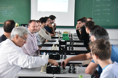 Fila de mesas com tabuleiros de xadrez. 14 pessoas aparecem jogando, de perfil, dos dois lados da foto. 
