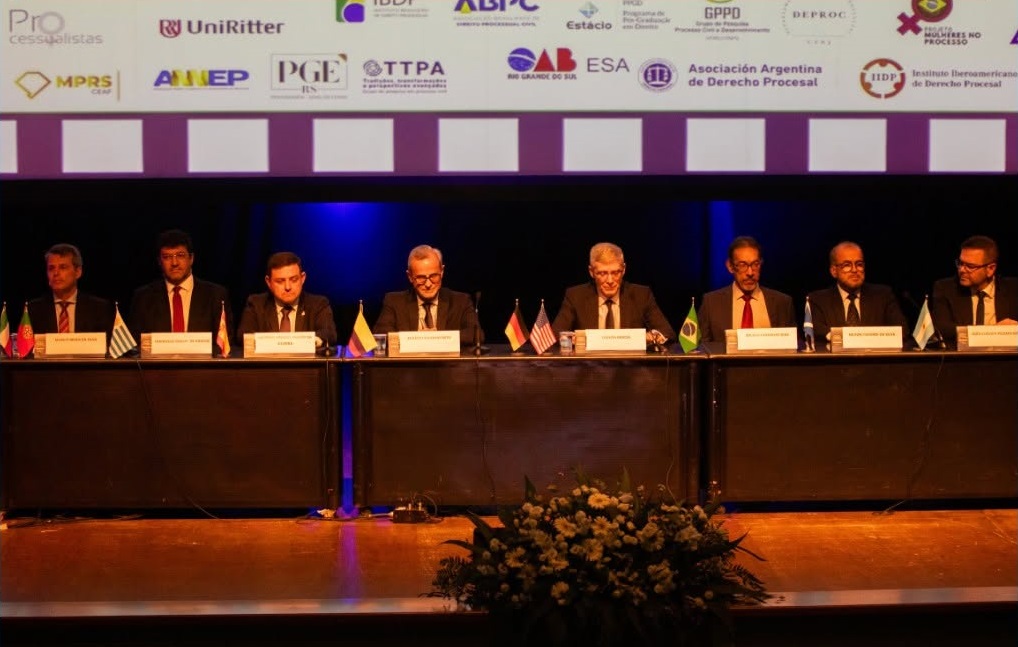 Mesa de abertura de um evento jurídico com oito homens trajando ternos sentados lado a lado, cada um com uma plaquinha de identificação e bandeiras de diferentes países à frente. Atrás deles, um telão exibe logotipos de instituições apoiadoras, como UniRitter, OAB-RS, MPRS, PGE-RS e outras. O ambiente é um auditório com iluminação cênica.