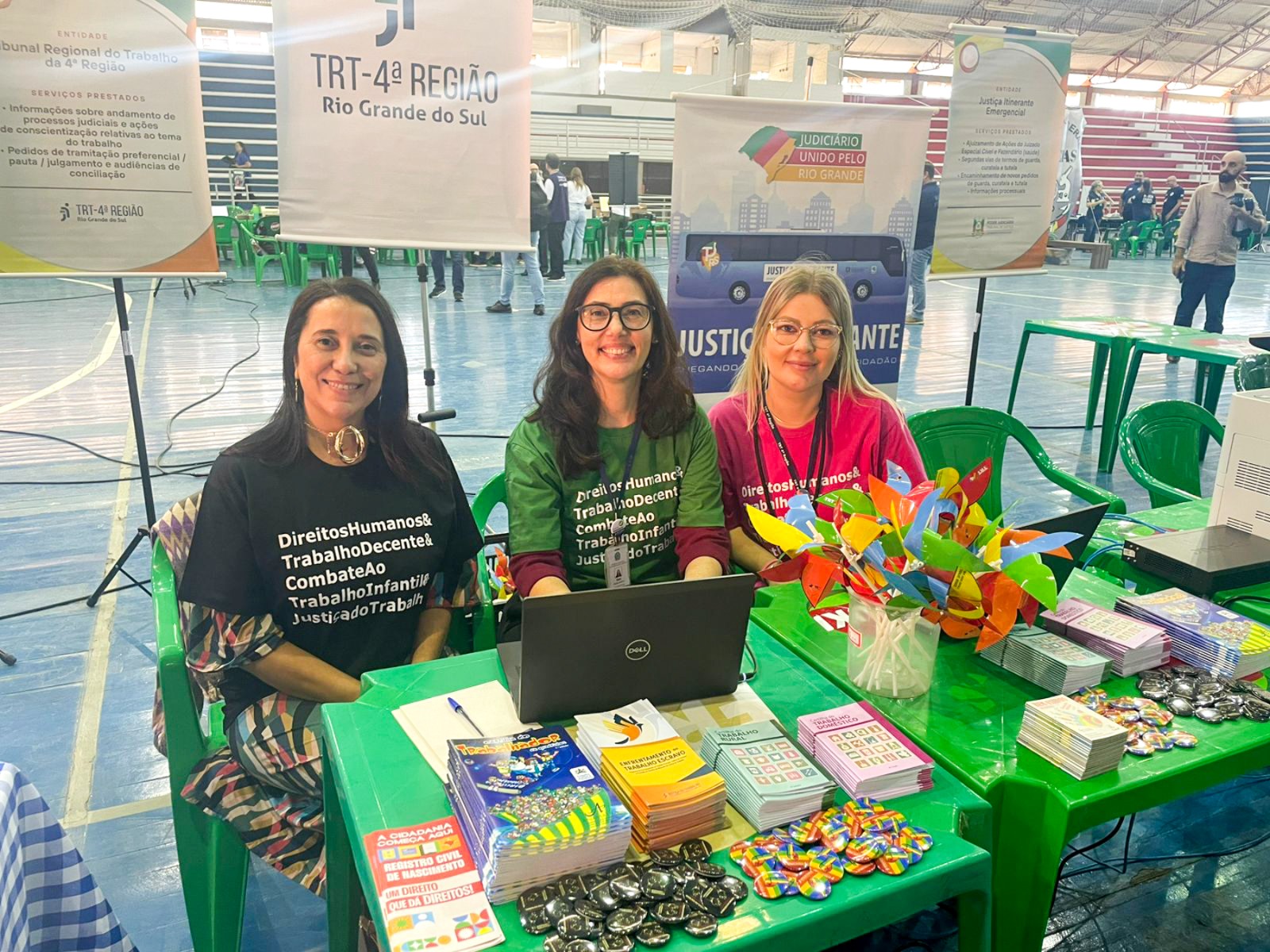 Três mulheres estão sentadas lado a lado atrás de uma mesa, sorrindo para a câmera. Elas usam camisetas com frases sobre direitos humanos e combate ao trabalho infantil. A mesa está repleta de materiais informativos e coloridos, como panfletos, livretos e cata-ventos de papel. Ao fundo, há banners com logotipos de órgãos públicos, dentro de um ginásio com arquibancadas.