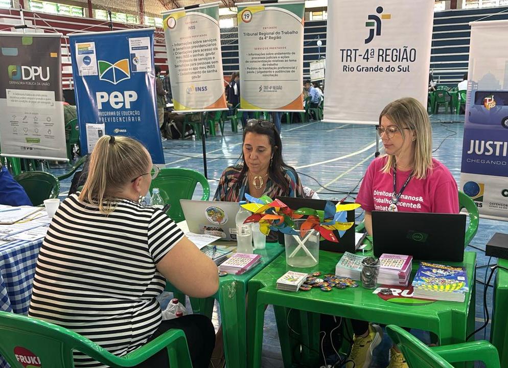Duas mulheres estão atendendo uma terceira, que está sentada do outro lado da mesa. Todas estão em um ginásio. A mesa é de plástico verde, com muitos materiais informativos e cata-ventos coloridos como decoração. Atrás, há diversos banners de instituições públicas, como o TRT-RS.