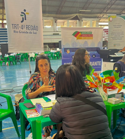 Uma mulher conversando em uma mesa de plástico verde, em um ginásio coberto, com cadeiras e mesas ao fundo. Há banners ao redor com os nomes de órgãos públicos, como o TRT-RS e o programa Justiça Itinerante. A mesa tem panfletos e pequenos brindes coloridos.