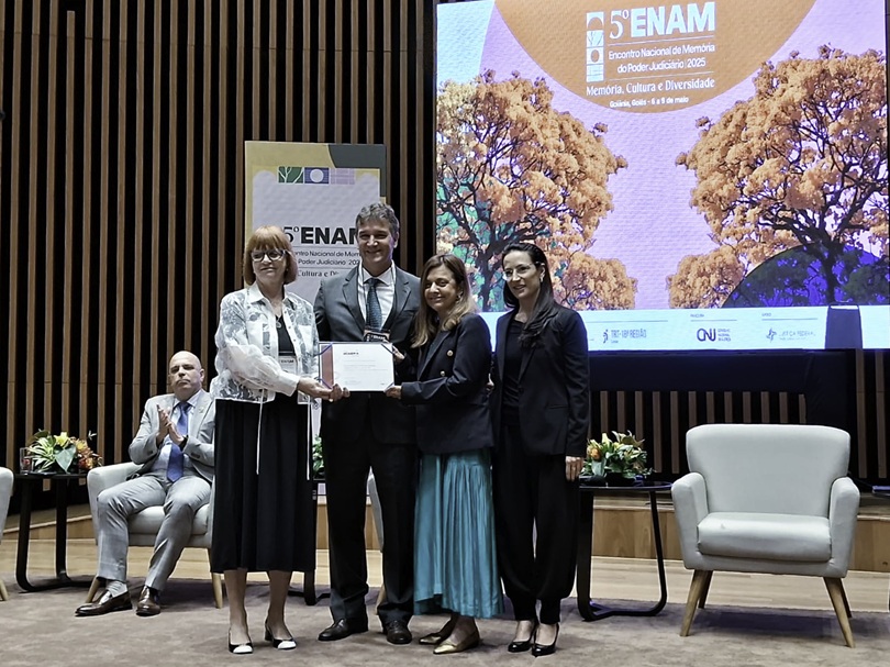 Cinco pessoas no palco do evento. Ao fundo aparecem um cartaz e um telão com a arte do Enam. As magistradas seguram o certificado da premiação e sorriem.