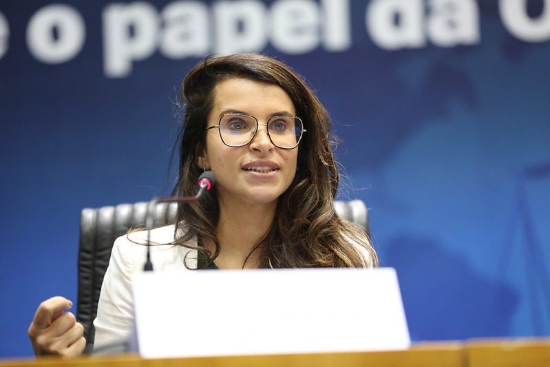 Carolina é branca, de cabelo liso castanho. Usa óculos de aro grande, fala ao microfone e gesticula. Veste um blazer branco.