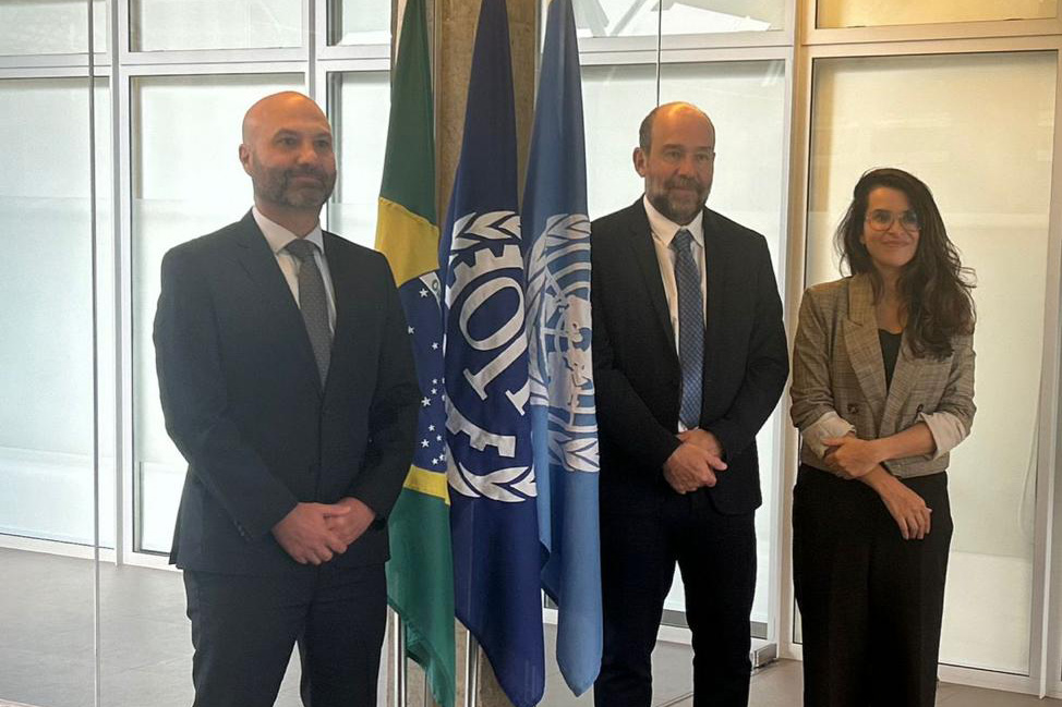 Foto posada, com as três pessoas de pé. Entre elas aparecem as bandeiras do Brasil, da OIT e da ONU.