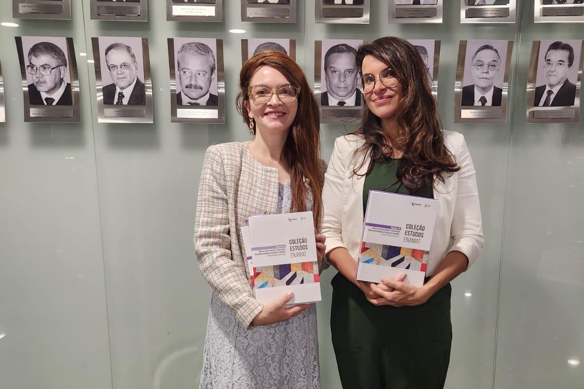 As duas magistradas posam para a foto segurando volumes da coleção estudos enamat. Ao fundo aparecem vários retratos em preto e branco na parede.