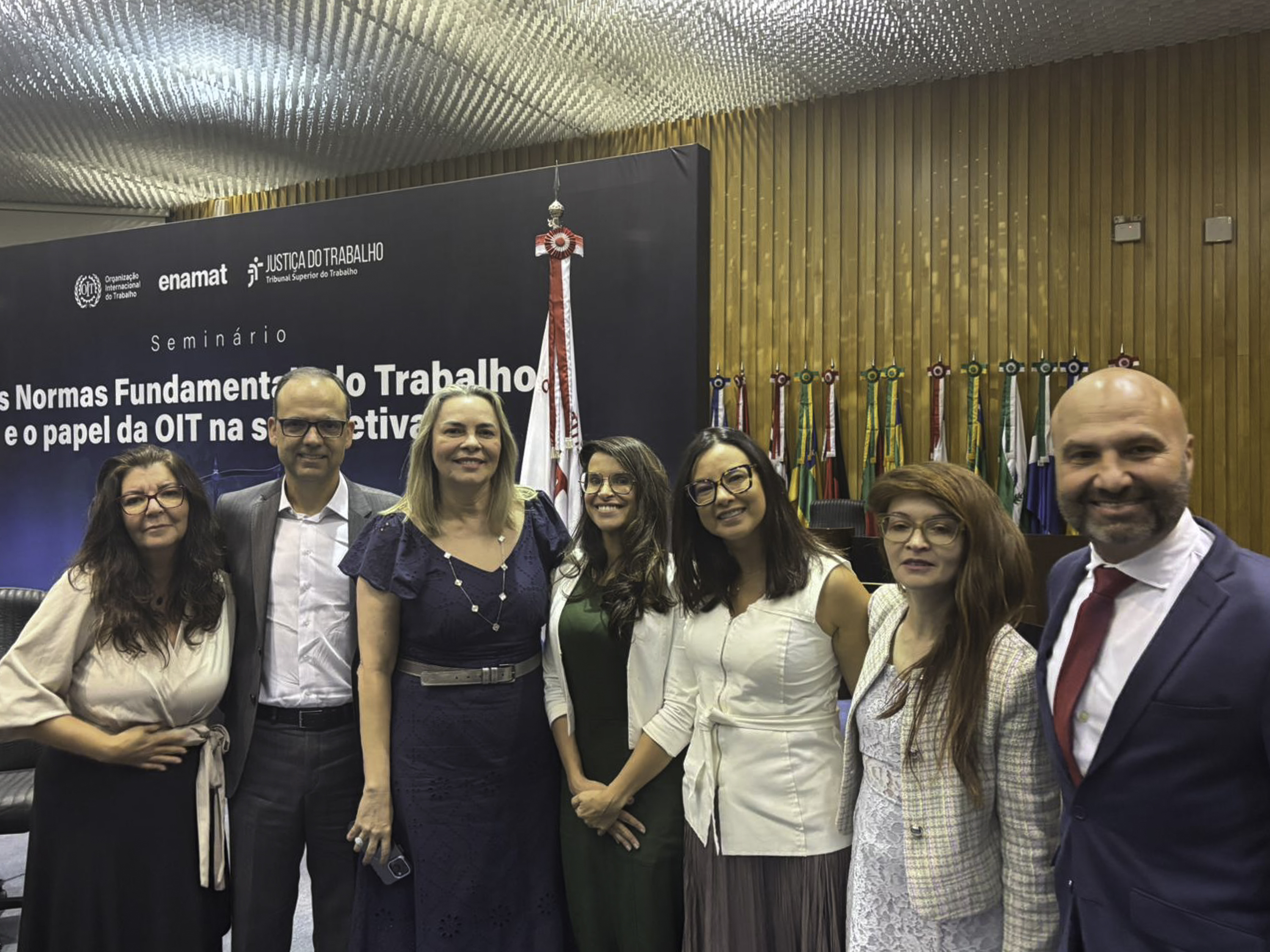 Sete pessoas posam para a foto. Ao fundo aparece um telão com o nome do seminário e diversas bandeiras