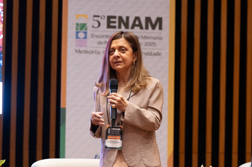Anita é branca, de cabelo escuro liso e comprido. Fala de pé ao microfone e usa blazer creme. Ao fundo aparece o logo do 5º Enam