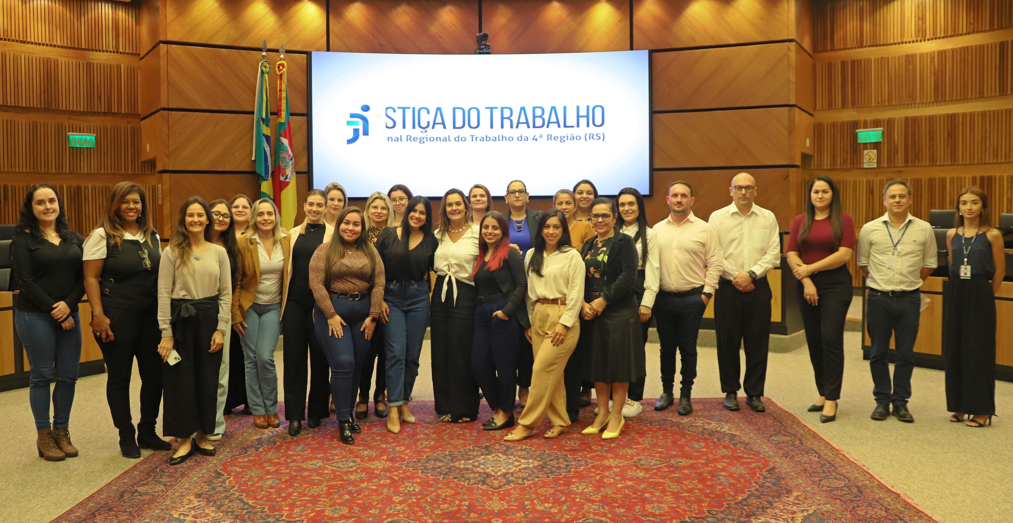 Grupo de estudantes de Direito da Ulbra Canoas posa para foto oficial no plenário do Tribunal Regional do Trabalho da 4ª Região (RS), ao lado da professora Juliana e da juíza auxiliar da Vice-Presidência, Luciana Xavier. Ao fundo, o telão exibe o logotipo da Justiça do Trabalho, e as bandeiras do Brasil, do Rio Grande do Sul e do TRT compõem o cenário institucional.