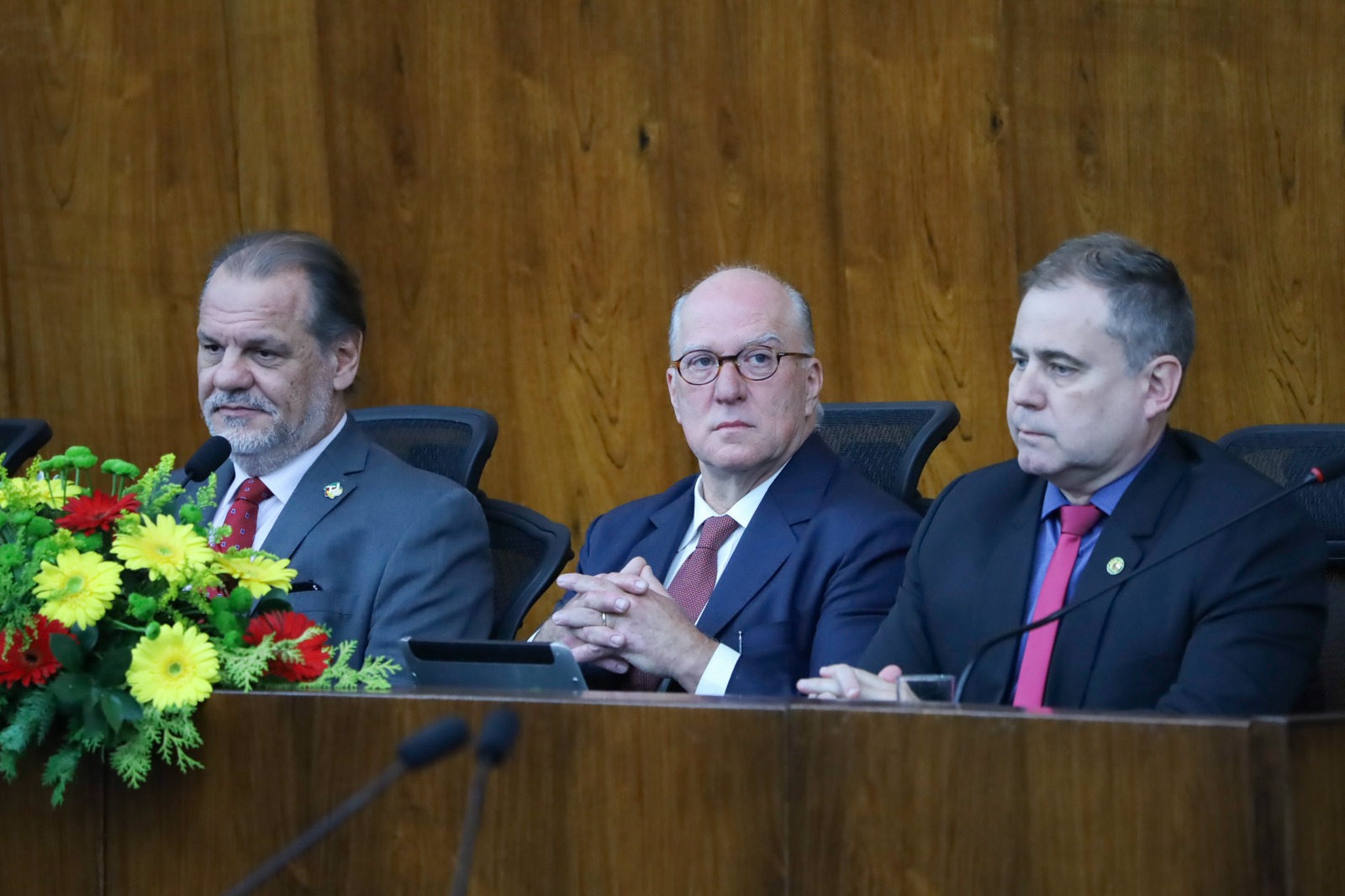 Três pessoas aparecem sentadas na mesa oficial. Na esquerda aparece um arranjo de flores.