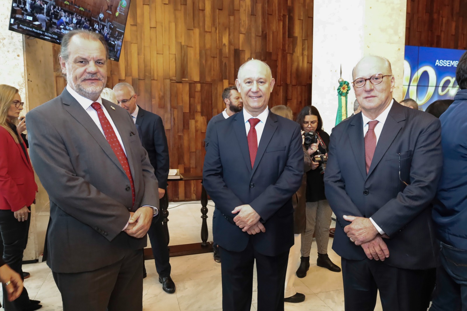 Três pessoas perfiladas posam para a foto. Eles usam terno e gravata e sorriem.
