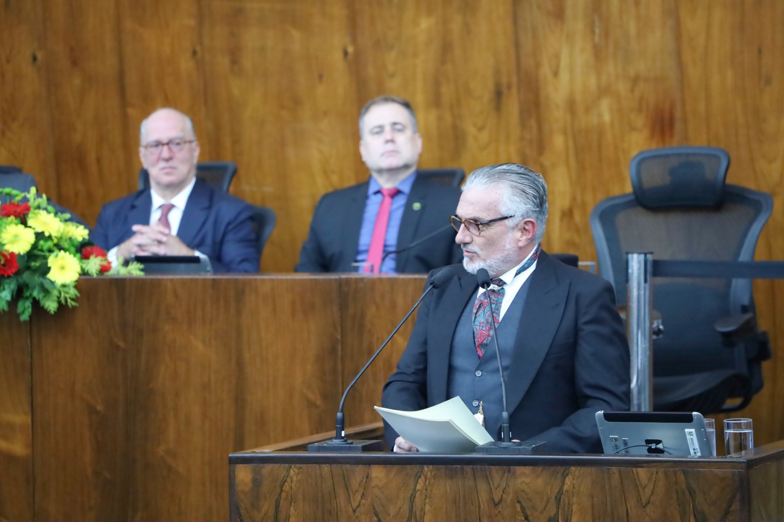 Home grisalho de barba aparece falando ao microfone. Ao fundo duas pessoas desfocadas aparecem na mesa oficial.