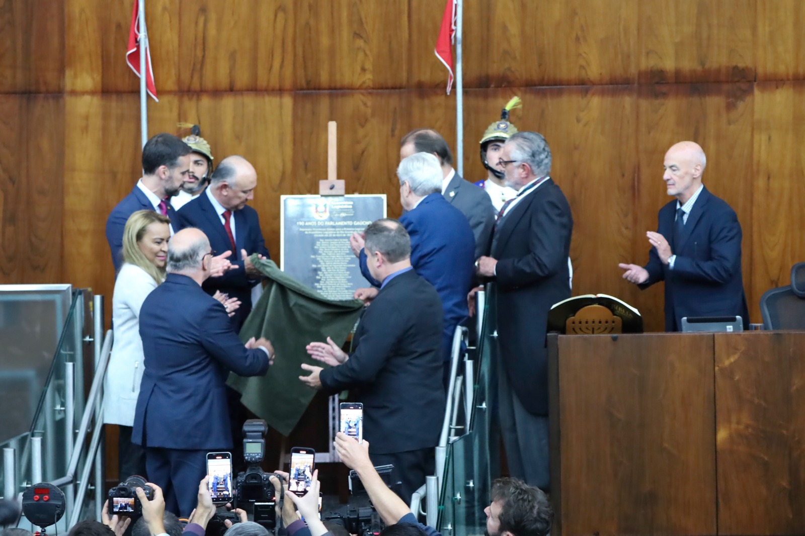 Pessoas aparecem em torno de um placa que é descerrada. Todos aplaudem.