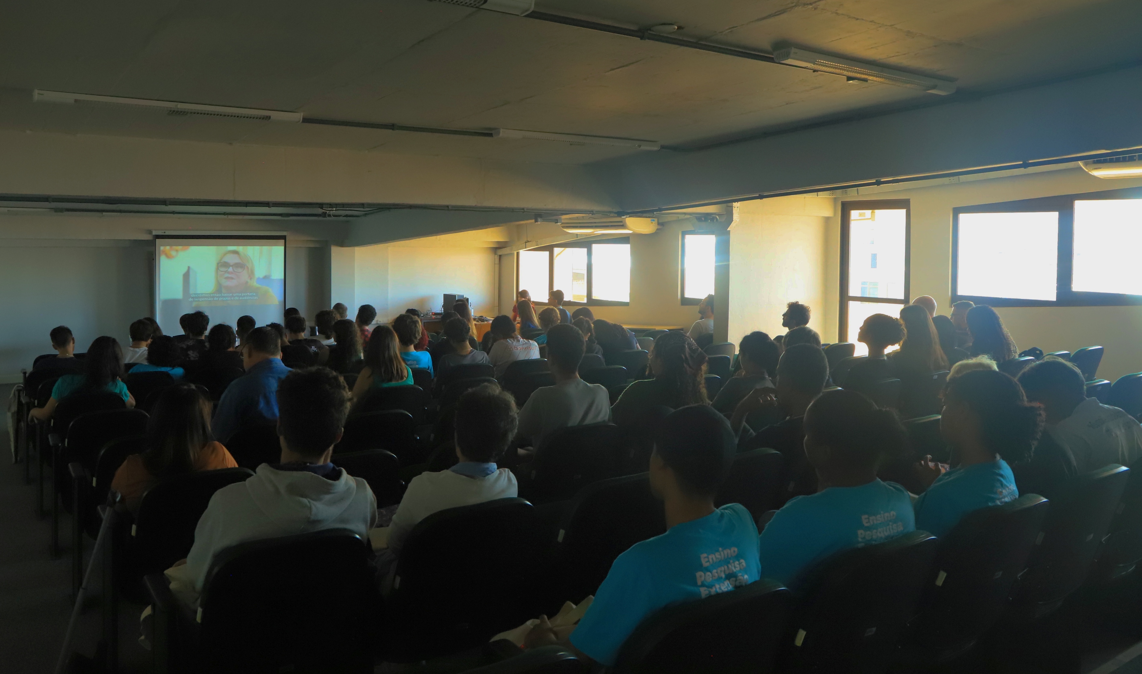 Alunos e professores do Instituto Federal do Rio Grande do Sul (IFRS) assistem, em um auditório escurecido, à exibição do documentário "A grande enchente de 2024 e a reconstrução da Justiça", produzido pelo Tribunal Regional do Trabalho do Rio Grande do Sul (TRT-RS). A projeção é exibida em uma tela grande à frente da sala, e alguns estudantes usam camisetas azuis com os dizeres “Ensino, Pesquisa e Extensão”.
