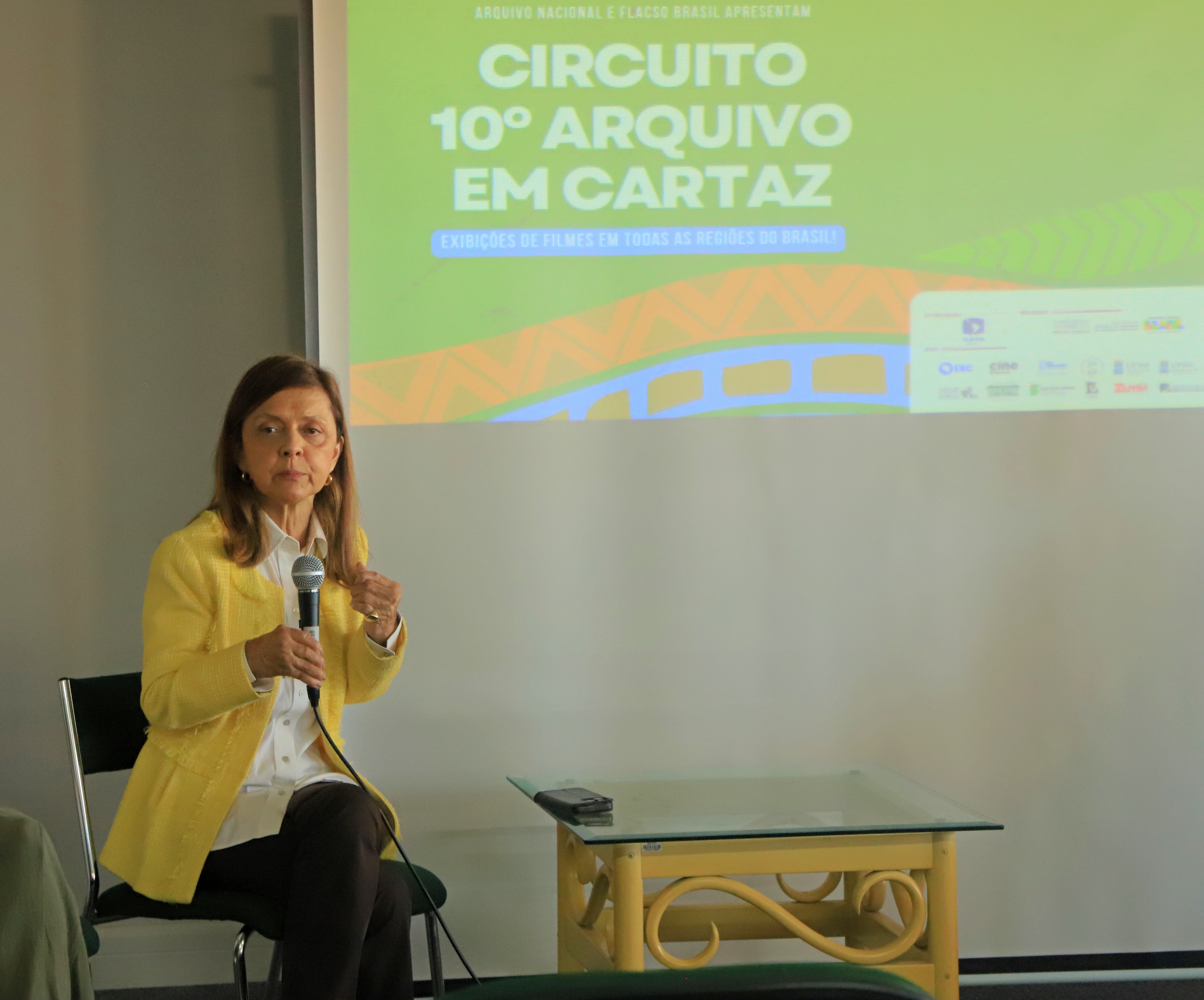 A juíza Anita Lübbe, do TRT-RS, participa de um bate-papo com os alunos após a exibição do documentário "A grande enchente de 2024 e a reconstrução da Justiça", no auditório do IFRS. Ela está sentada, segurando um microfone, diante de uma tela que exibe o cartaz do evento “Circuito 10º Arquivo em Cartaz – Exibições de filmes em todas as regiões do Brasil!”. Veste blazer amarelo claro e camisa branca.