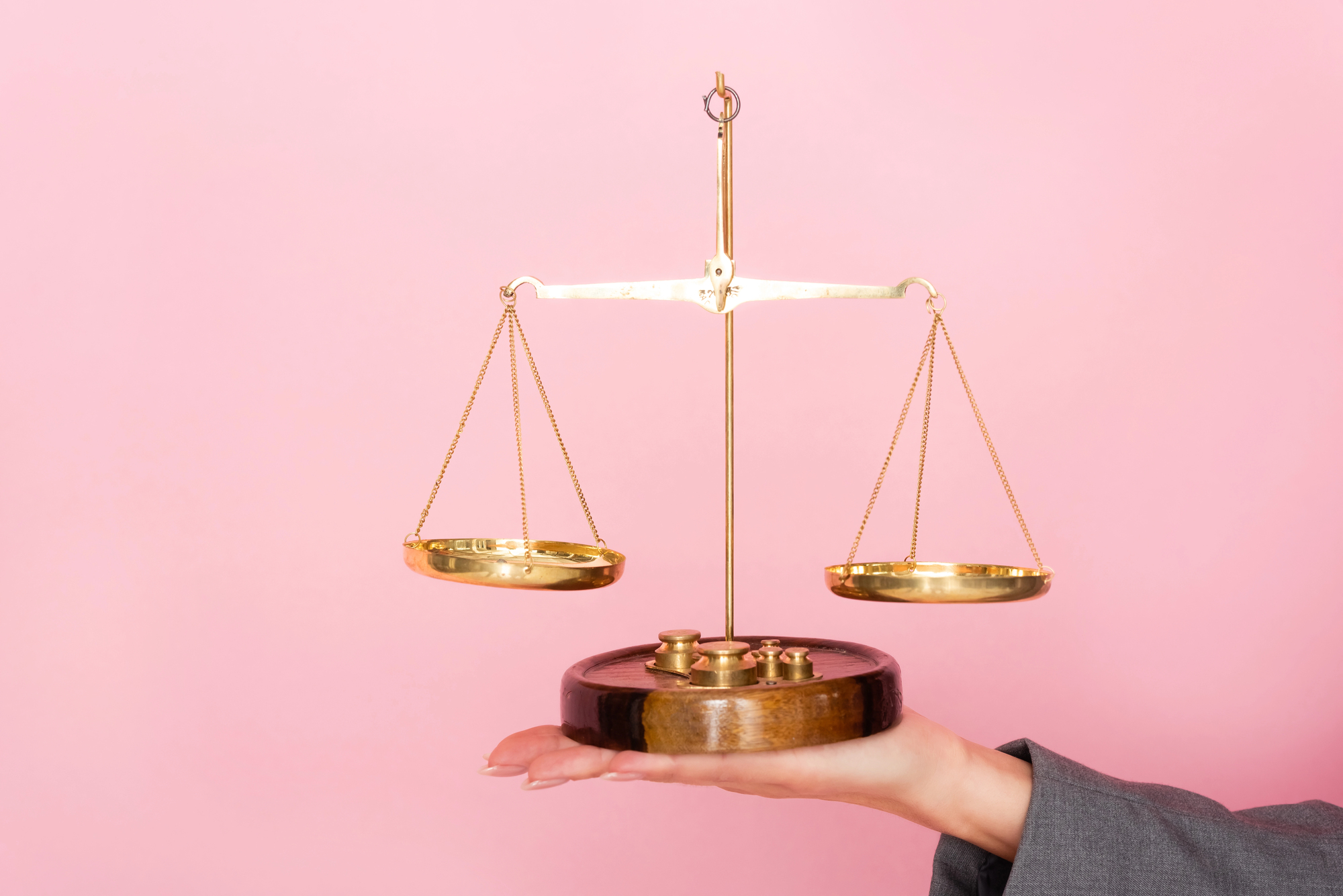 Balança de ouro desequilibrada, com base de madeira escura, sendo segurada por uma mão feminina com unhas bem feitas. O fundo é rosa claro, transmitindo uma estética suave e minimalista.