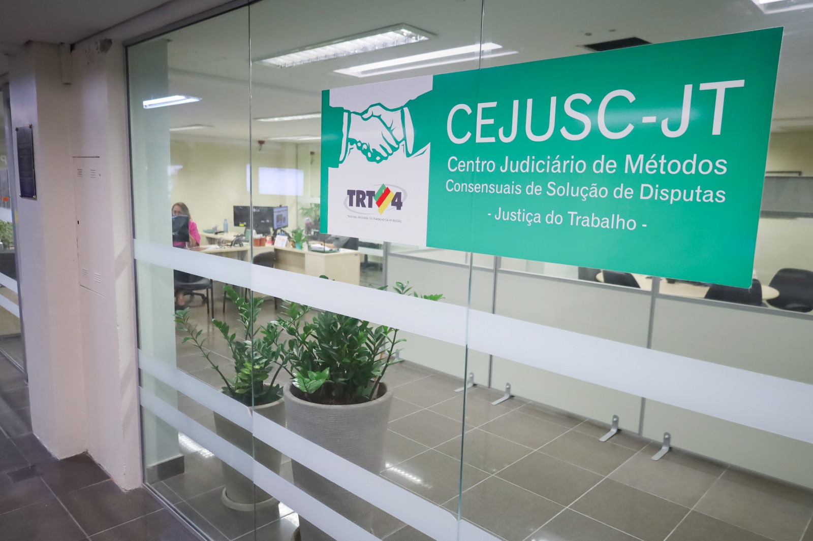 Fachada envidraçada de uma sala. No vidro da porta há um cartaz com a sigla Cejusc-JT e o nome da unidade por extenso. No lado esquerdo há uma arte com o desenho de um aperto de mão sobre o logo do TRT-4.