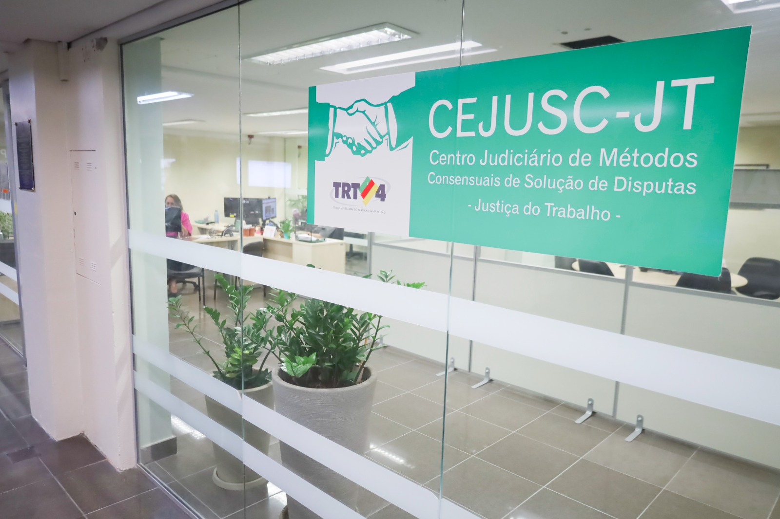 Fachada envidraçada de uma sala. No vidro da porta há um cartaz com a sigla Cejusc-JT e o nome da unidade por extenso. No lado esquerdo há uma arte com o desenho de um aperto de mão sobre o logo do TRT-4.