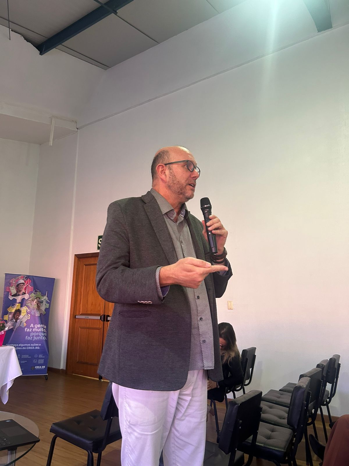 Um homem em pé, posicionado de frente para a câmera. Ele está usando uma camisa clara e palestrando.