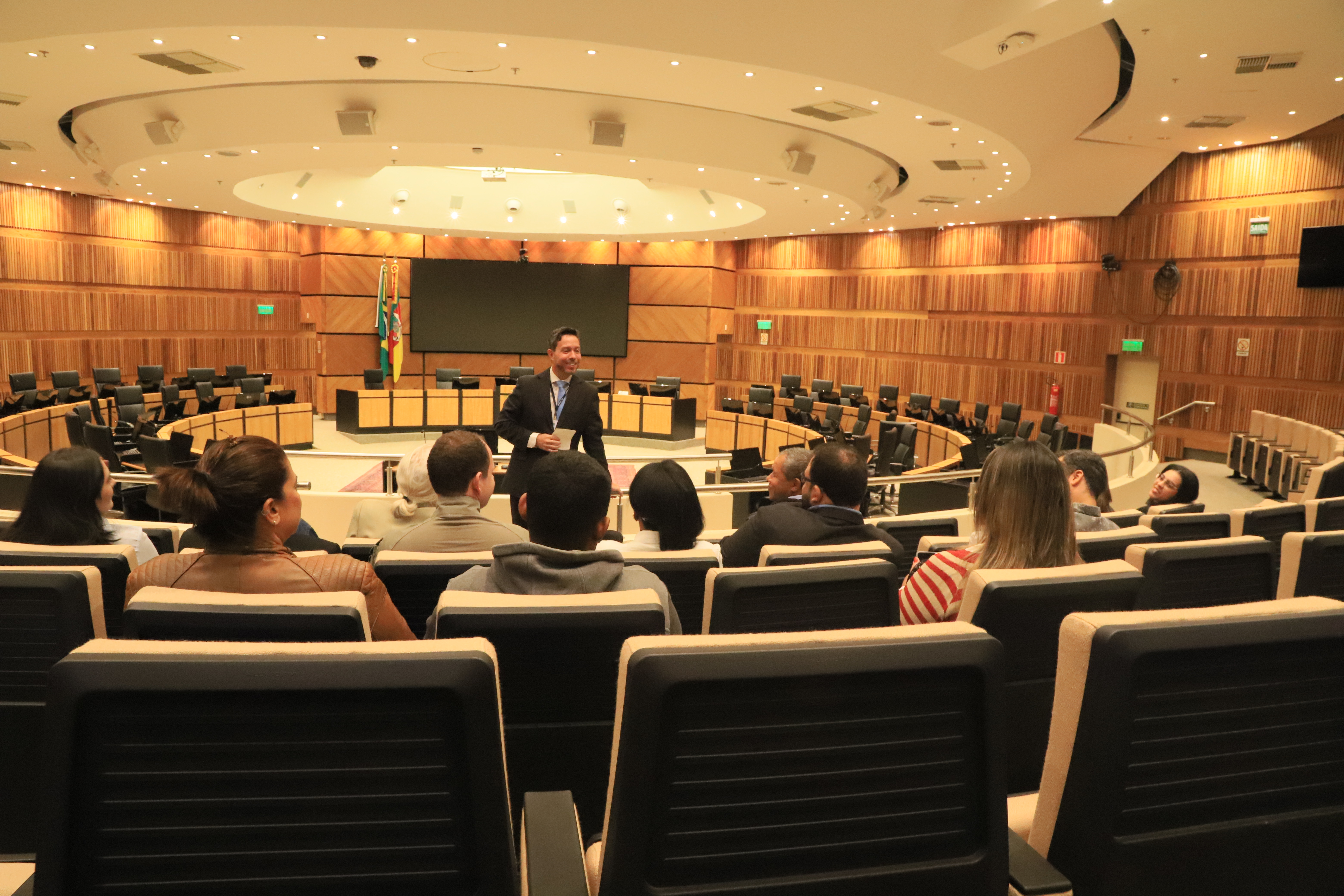 Juiz Rodrigo Trindade conversa com alunos(as) e professores(as) no Plenário Milton Varela, em frente ao painel e bandeiras, em ambiente moderno e iluminado.