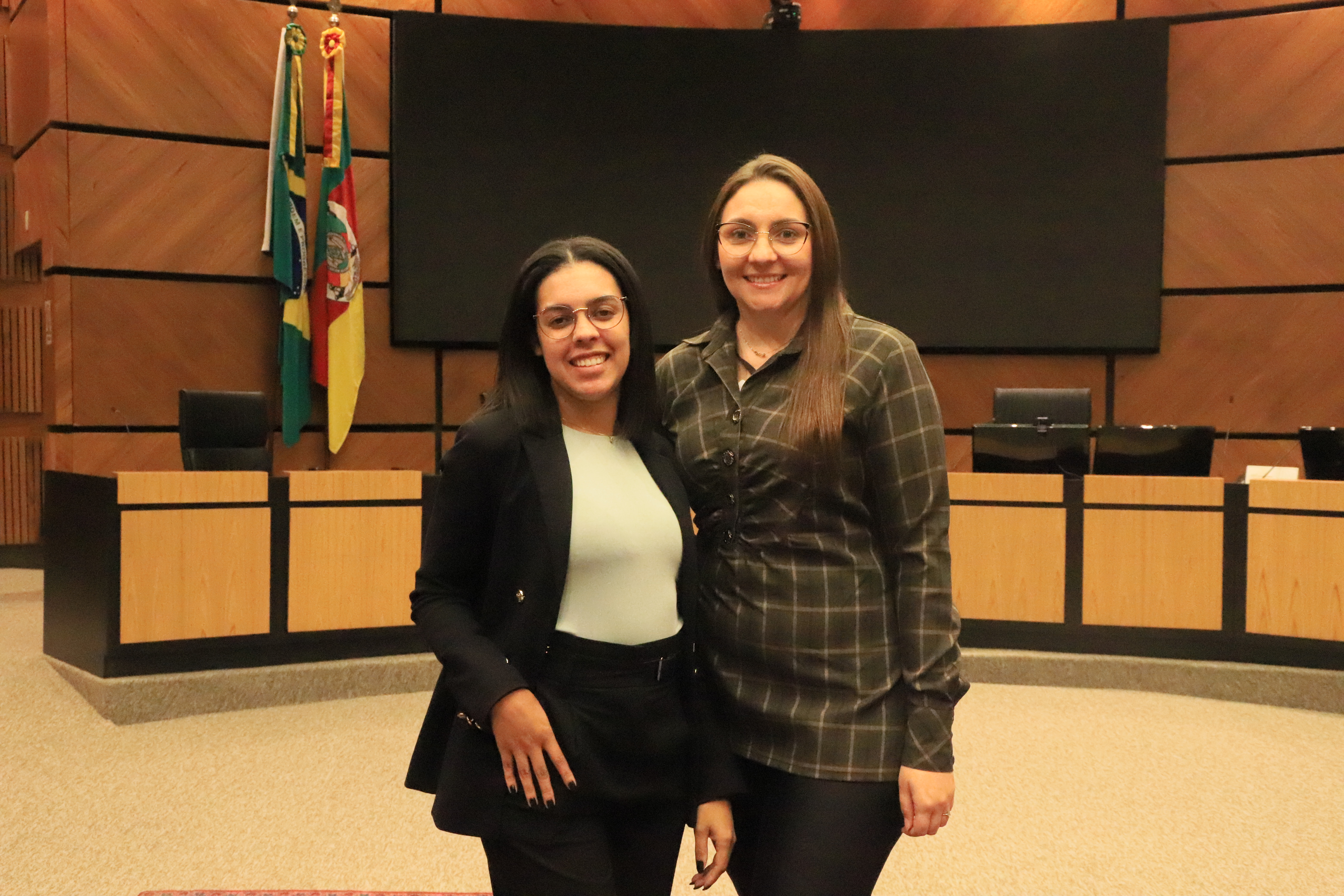 Duas alunas posam sorridentes em frente ao plenário Milton Varela, com bandeiras ao fundo e telão desligado.