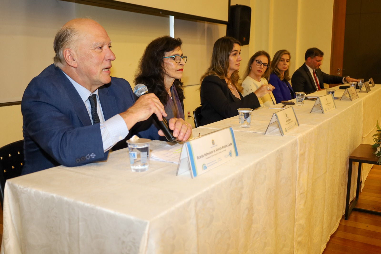 A imagem mostra o presidente Martins Costa discursando na mesa de autoridades.