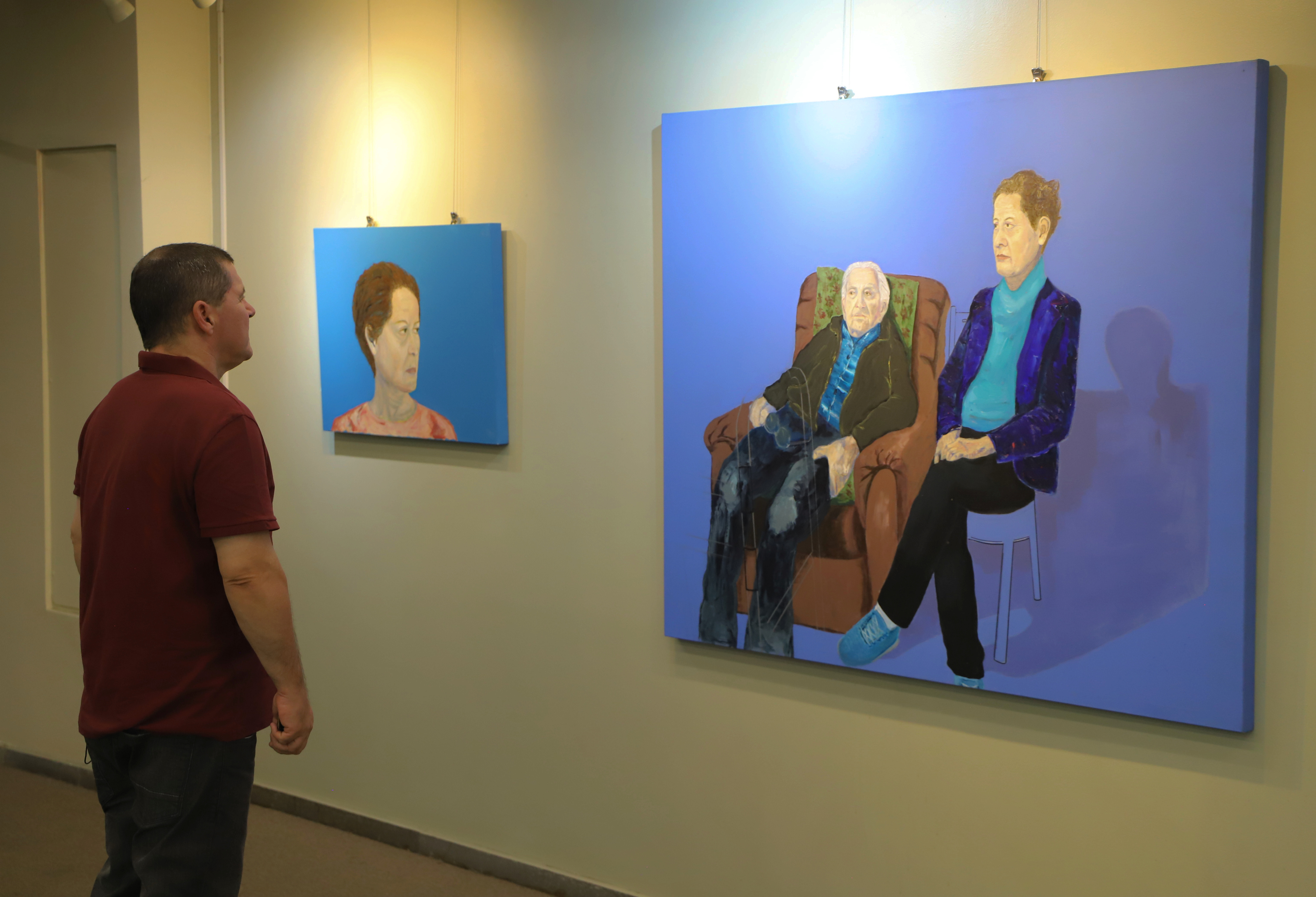 Homem observa duas pinturas expostas em uma galeria de arte. A primeira, menor, retrata o rosto de uma mulher com expressão séria sobre fundo azul. A segunda, maior, mostra dois idosos sentados lado a lado — um homem em uma poltrona marrom e uma mulher em um banco — ambos também em frente a um fundo azul. A iluminação destaca as obras em contraste com a parede clara da galeria.