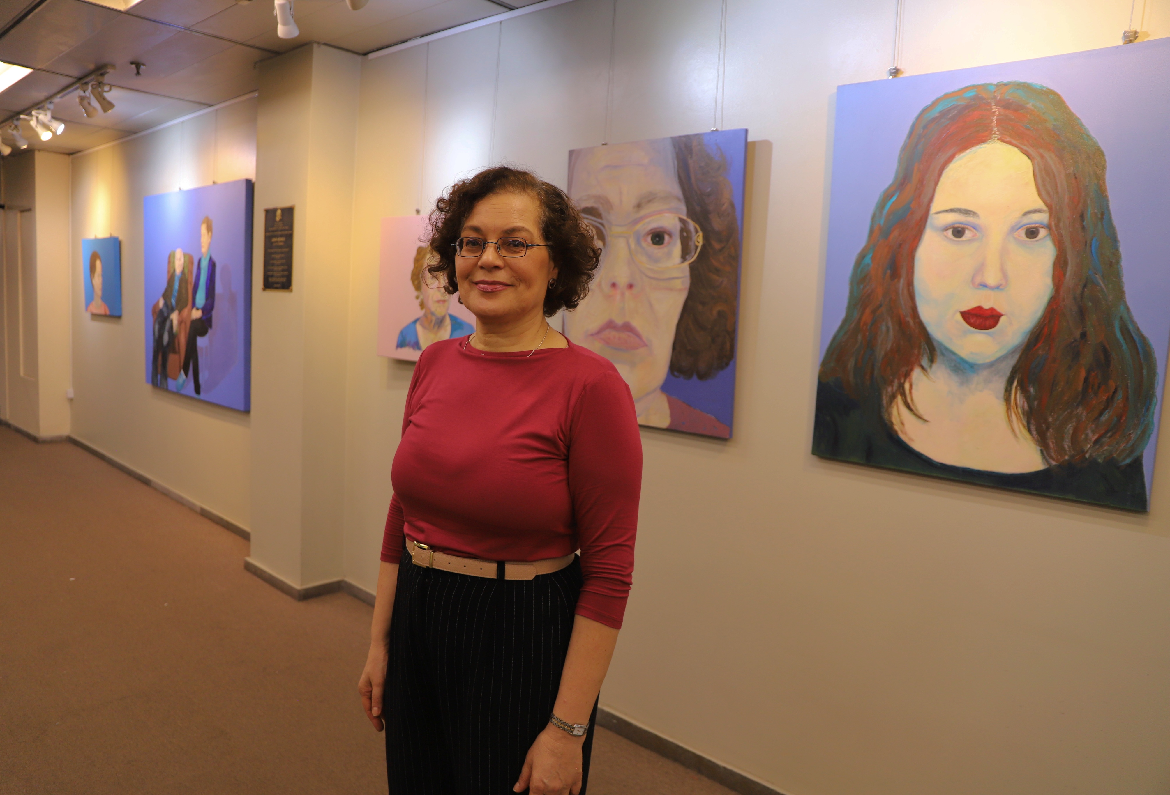 Mulher de cabelos cacheados e curtos, vestindo blusa rosa e calça escura com cinto bege, posa sorrindo em uma galeria de arte. Ao fundo, várias pinturas em estilo retratista estão expostas em uma parede clara. As obras mostram rostos femininos com expressões variadas sobre fundos coloridos, predominando tons de azul e roxo.