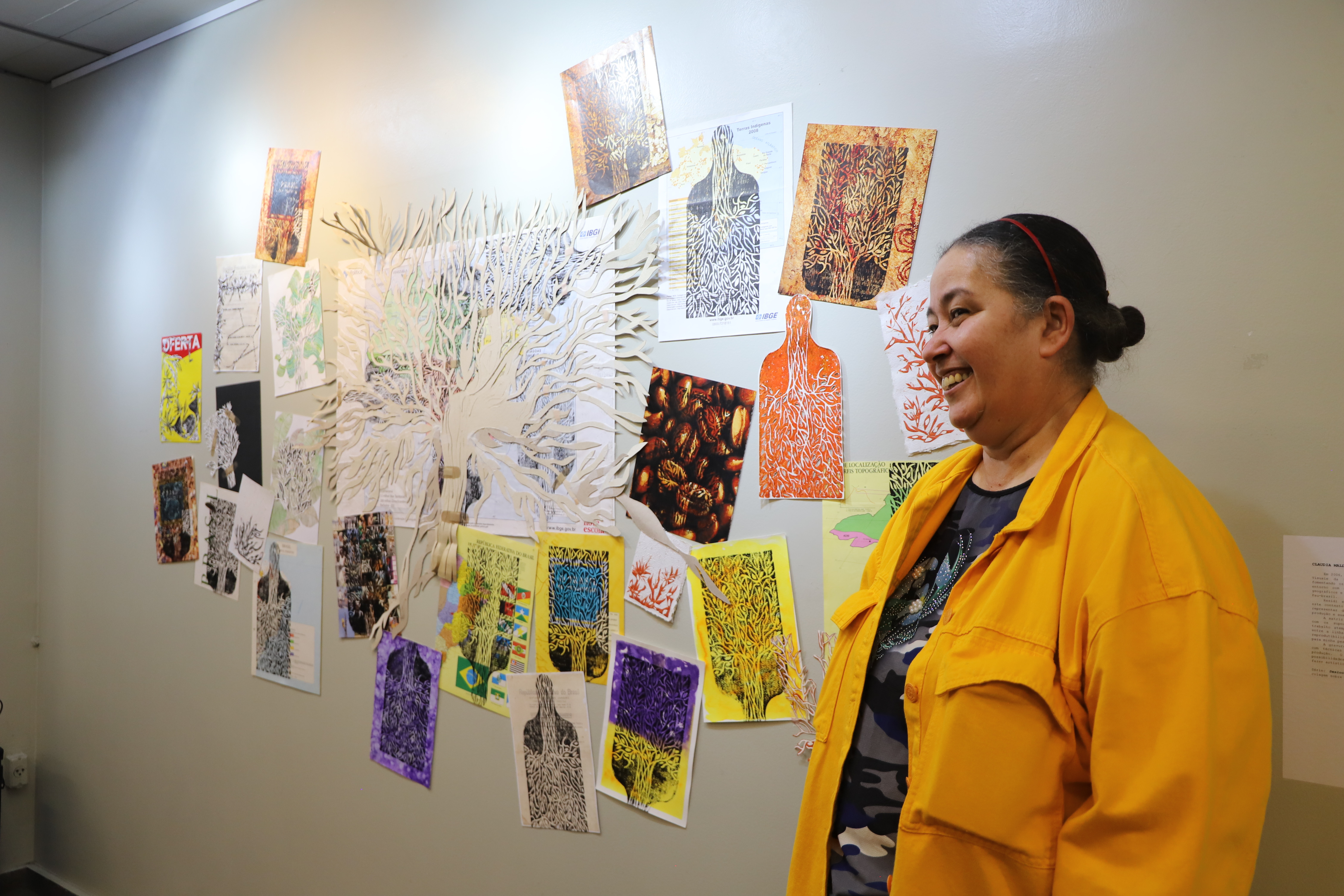 Mulher sorridente com os cabelos presos e vestindo uma jaqueta amarela posa ao lado de uma parede decorada com colagens artísticas. A instalação exibe recortes de figuras humanas preenchidas por padrões semelhantes a raízes ou galhos, em diversas cores, além de texturas e elementos orgânicos sobrepostos. A composição inclui papéis recortados, desenhos e colagens, criando uma obra visualmente rica e expressiva.