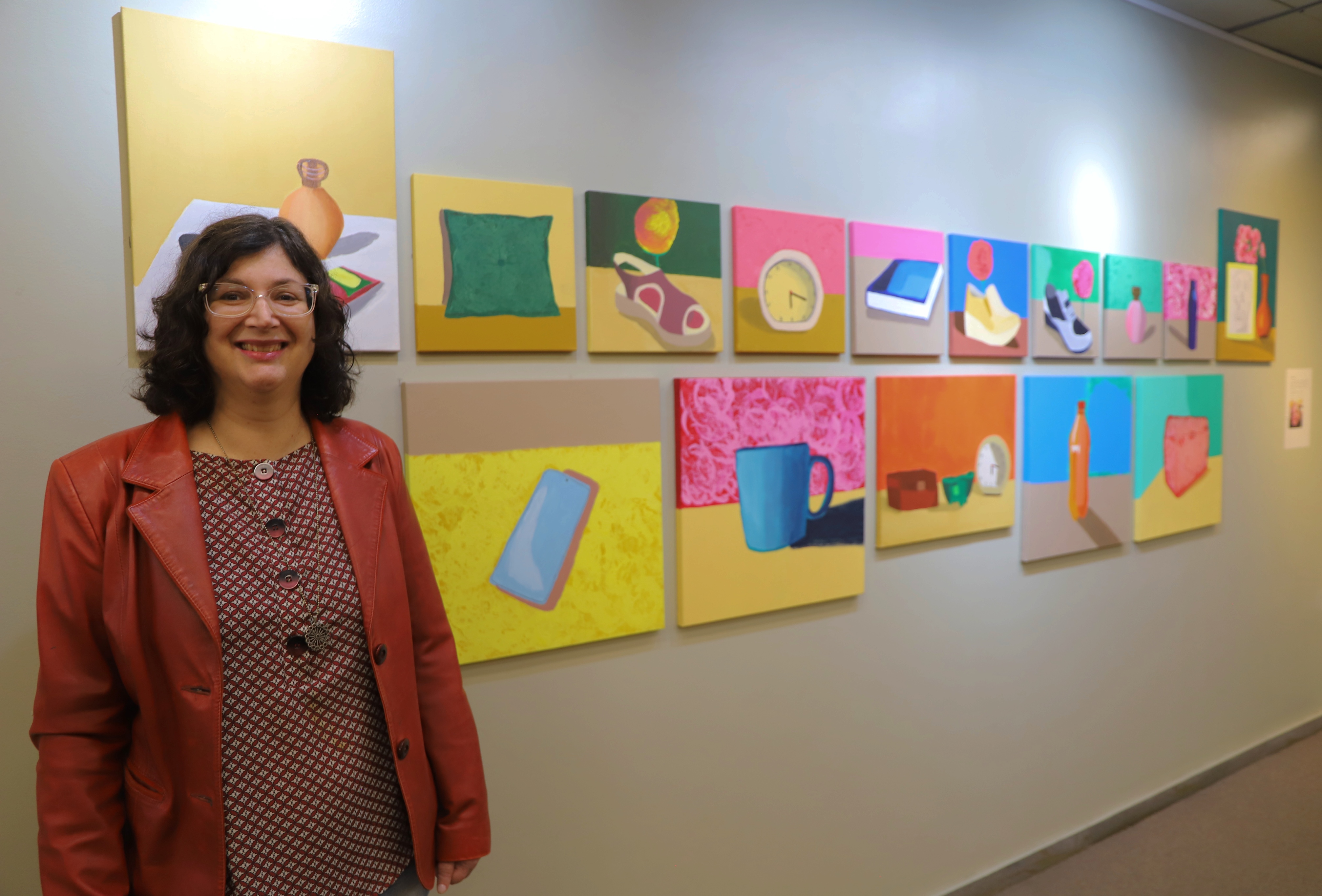 Mulher de cabelos ondulados, óculos e usando um blazer vermelho sobre uma blusa estampada sorri em frente a uma parede com uma exposição de quadros coloridos. As obras retratam objetos do cotidiano, como almofadas, sapatos, relógios, canecas e garrafas, pintados em estilo pop com cores vibrantes e fundos contrastantes. A disposição simétrica das telas em duas fileiras cria uma composição visual alegre e harmoniosa.