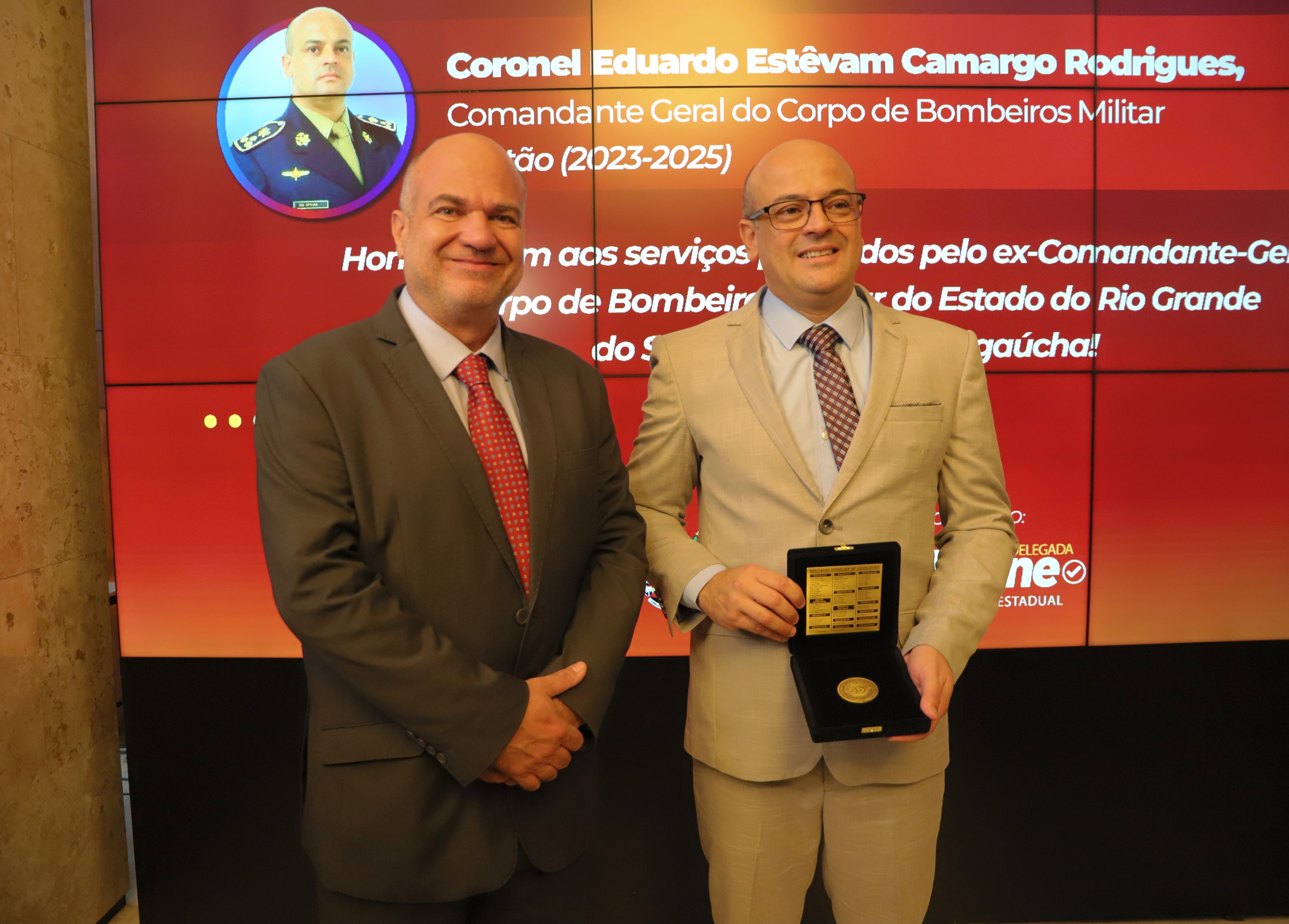 O desembargador Lucena, de terno cinza escuro e gravata vermelha, posa ao lado do oficial Eduardo Estêvam Camargo Rodrigues, que veste terno bege claro com gravata estampada e segura uma caixa aberta com uma medalha dourada. Ao fundo, um painel digital exibe o nome completo do agente, sua função como Comandante do Corpo de Bombeiros Militar e o período da gestão (2023–2025), além de uma mensagem de homenagem pelos serviços prestados.