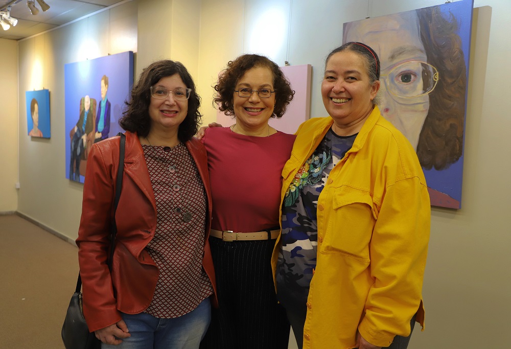 Três artistas em pé pousando para a foto em frente aos quadros expostos na parede