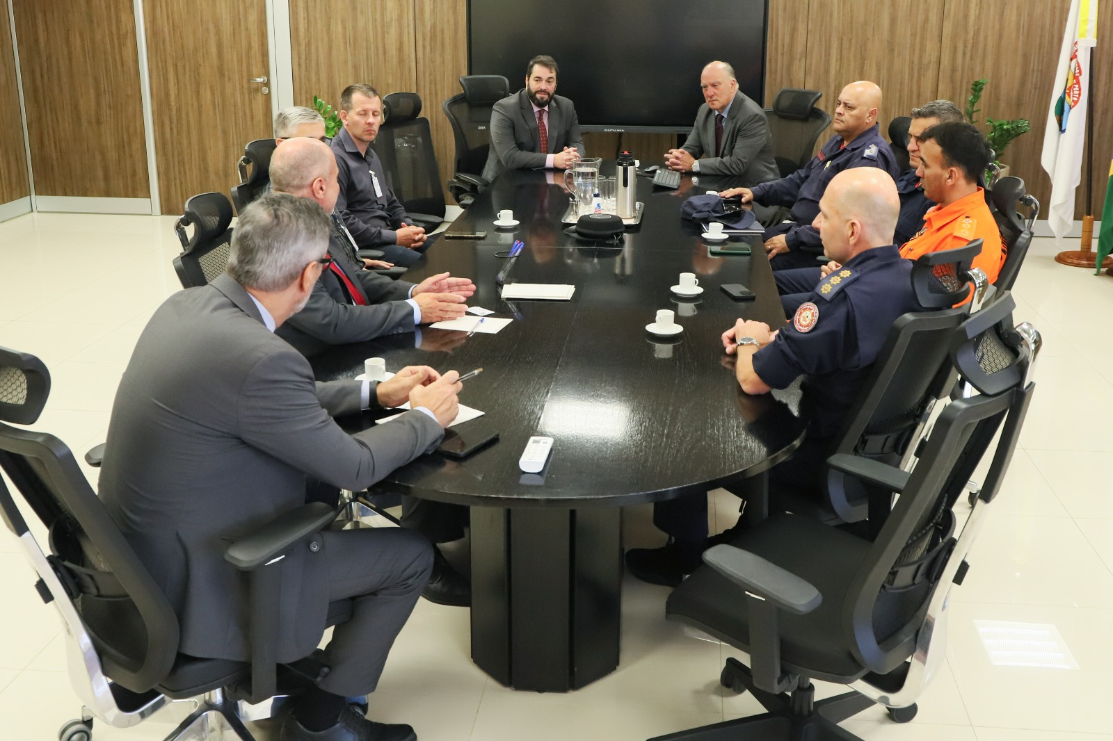 Participantes na mesa de reunião no Salão Nobre. Dez pessoas aparecem na imagem, com uma bandeira ao fundo.