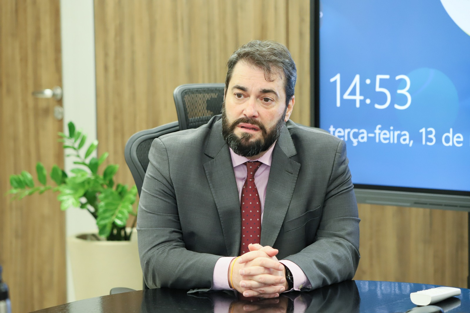 Alexandre Correa da Cruz é branco, tem cabelo escuro, usa terno escuro com camisa rosa e gravata bordô. Está com as mãos cruzadas sobre a mesa olhando para a esquerda da imagem.