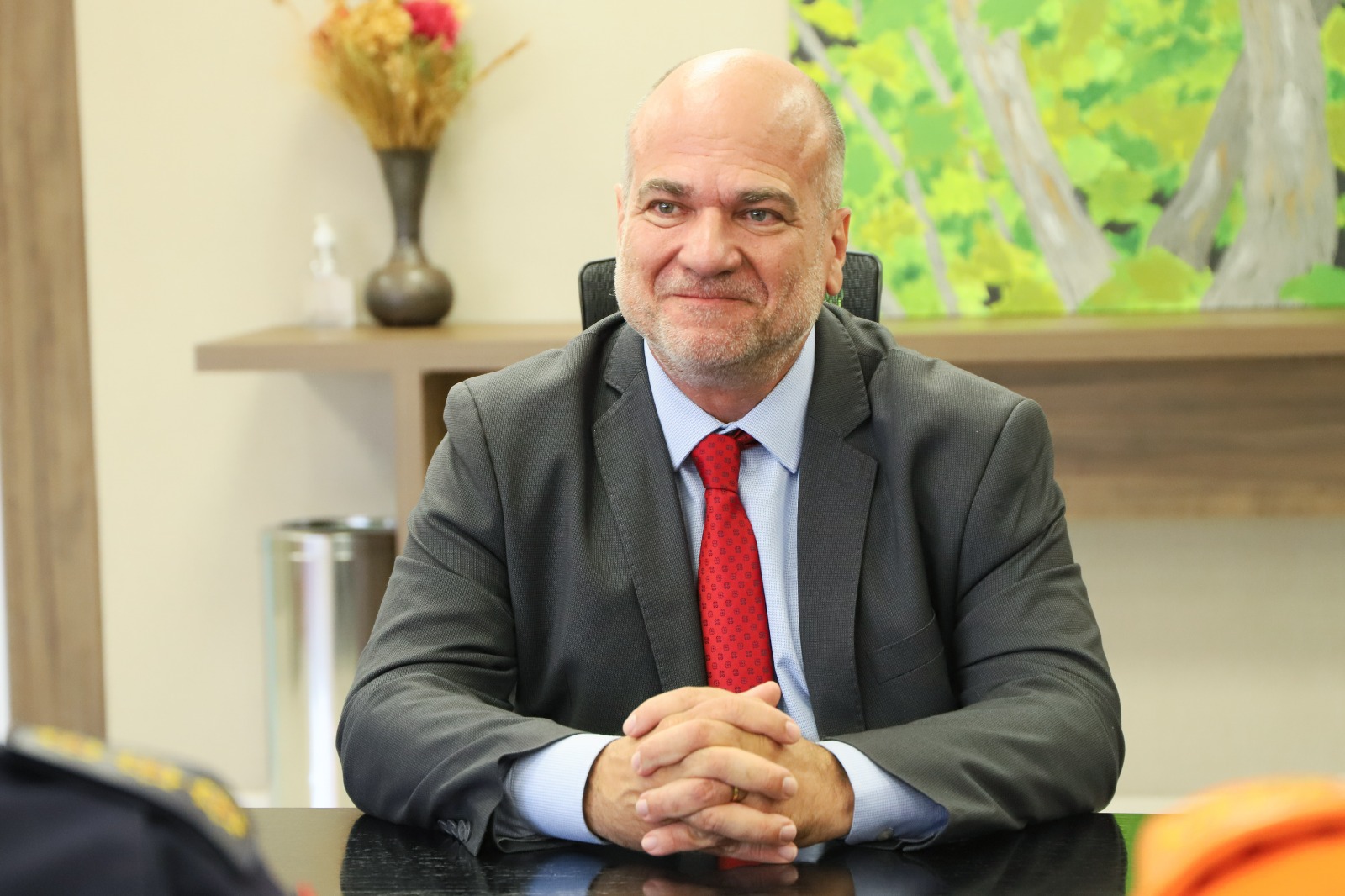 Lucena é branco, calvo, tem olhos claros. Veste terno cinza escuro, camisa branca e gravata vermelha. Está com as mãos cruzadas sobre a mesa e sorrindo.