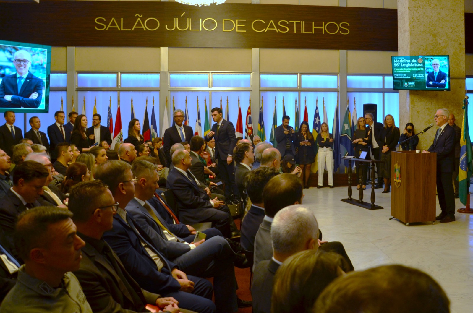 Folto em plano aberto da cerimônia na Assembleia, mostrando público sentado e procurador-geral falando no púlpito.