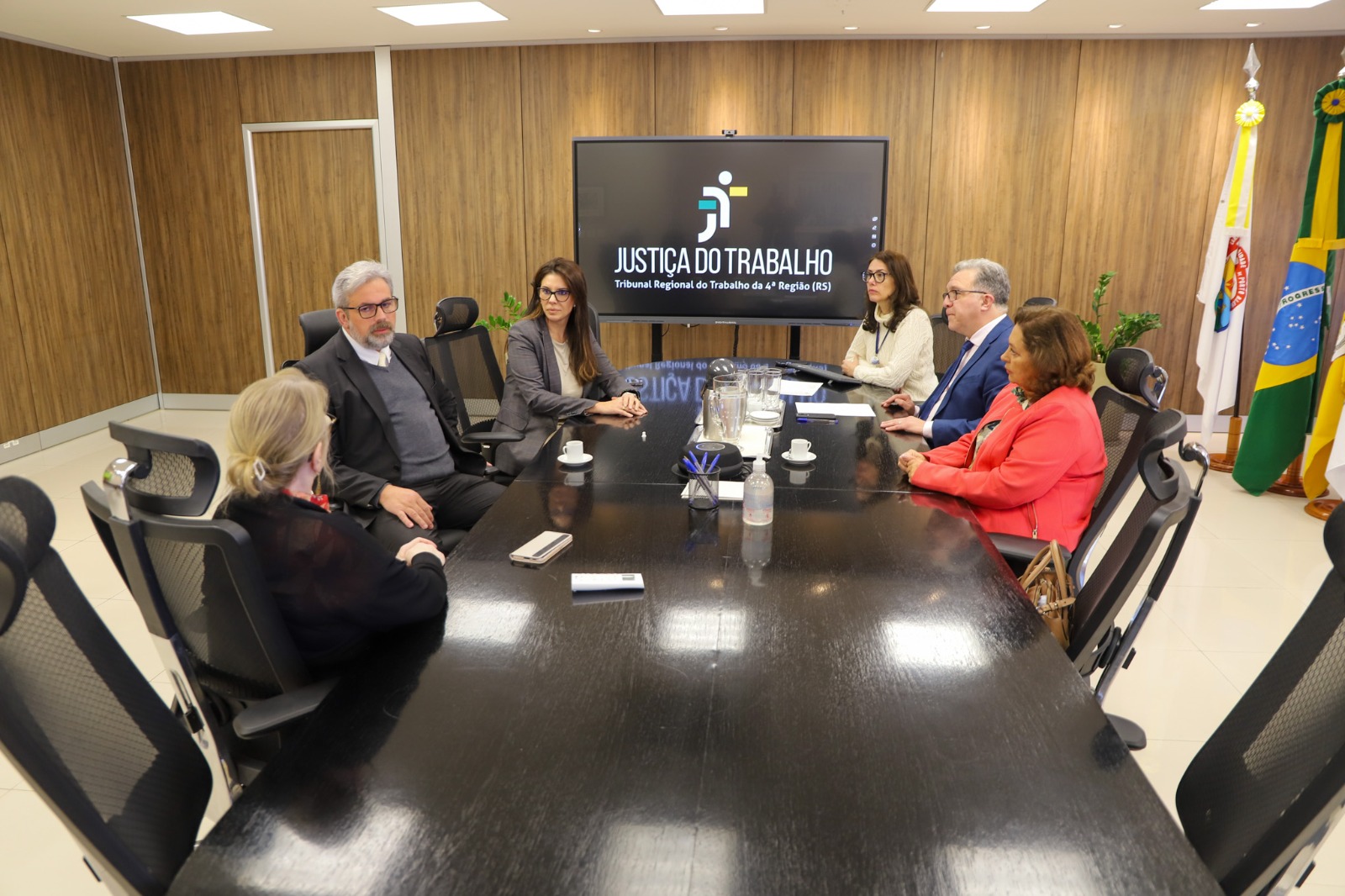 A foto mostra pessoas sentadas ao redor de uma mesa, numa reunião, com telão de fundo, escrito Justiça do Trabalho.