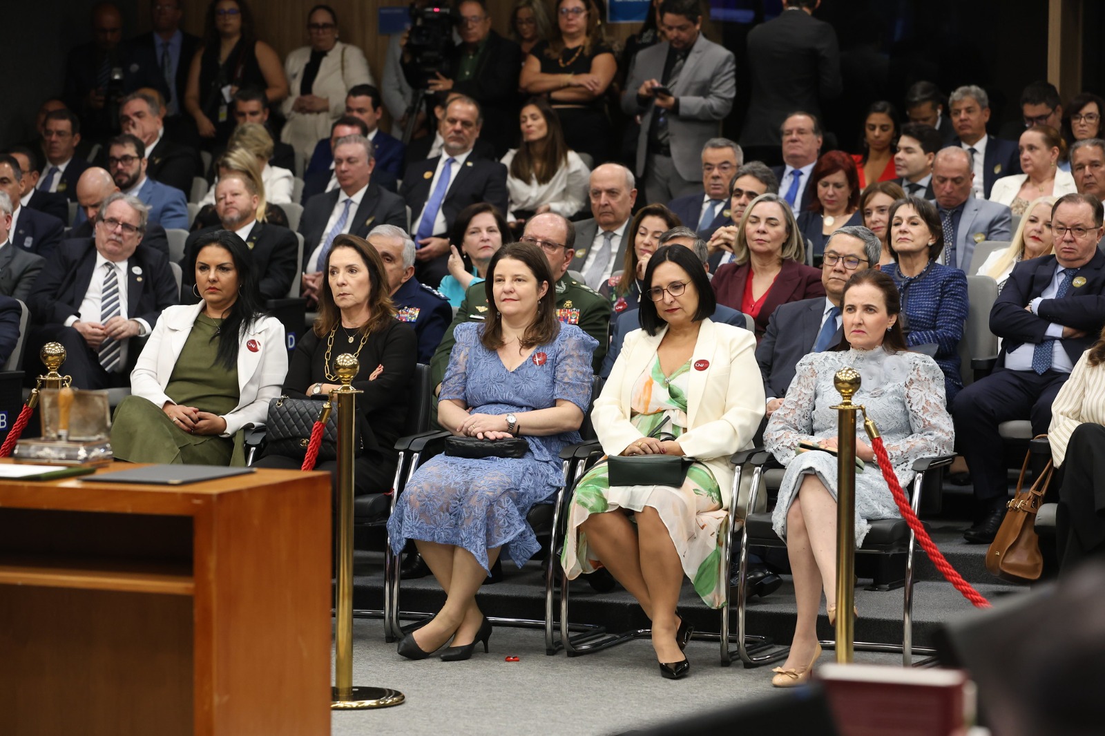 Público presente no plenário do Conselho Nacional de Justiça (CNJ), durante sessão solene. Em primeiro plano, mulheres sentadas nas primeiras fileiras, vestindo trajes sociais formais, algumas com vestidos e blazers claros. Atrás delas, várias autoridades, incluindo homens e mulheres, assistem atentos à cerimônia.