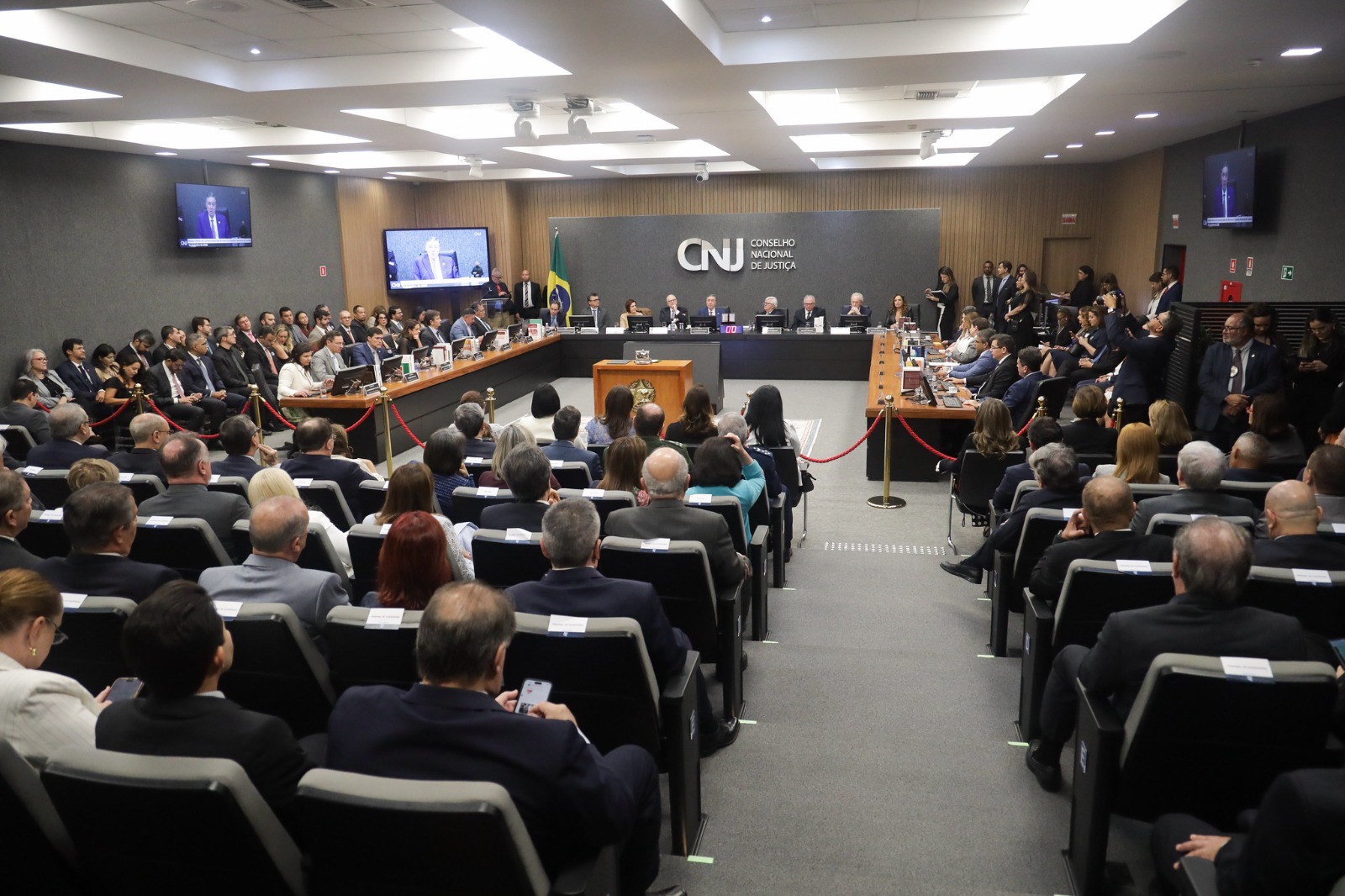 Visão geral do plenário do Conselho Nacional de Justiça (CNJ) durante sessão solene. A mesa diretora está posicionada ao fundo, com o logotipo do CNJ na parede e a bandeira do Brasil ao lado. O auditório está lotado, com o público sentado em fileiras de cadeiras cinza, enquanto jornalistas e servidores acompanham em pé nas laterais. Dois monitores de televisão exibem a transmissão ao vivo da sessão. Caption