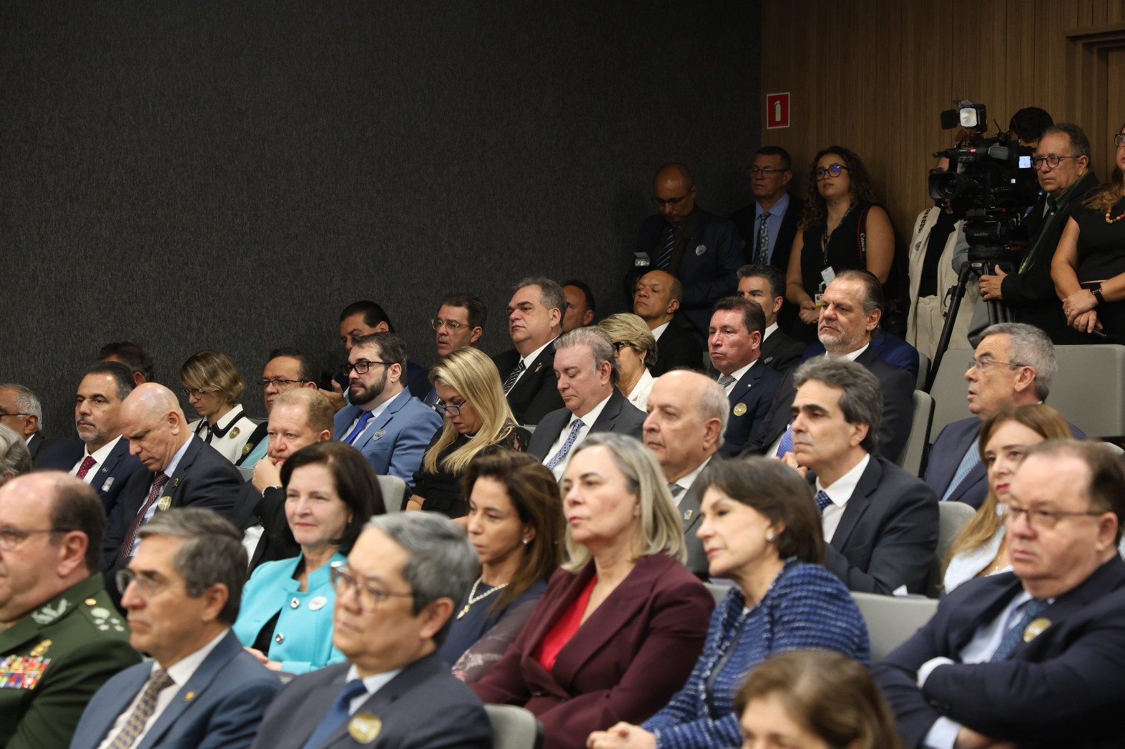 Autoridades e convidados acompanham atentamente a sessão solene no plenário do Conselho Nacional de Justiça (CNJ). A imagem mostra o público sentado em fileiras, com homens e mulheres vestidos formalmente, incluindo membros das Forças Armadas, magistrados e servidores. Ao fundo, alguns profissionais de imprensa registram o evento com câmeras.